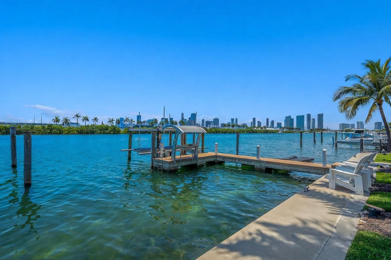 This scenic waterfront view captures a private wooden dock extending into a vibrant turquoise bay, complete with a boat lift and a clear view of the distant city skyline. The foreground features a concrete walkway lined with white lounge chairs, set against a backdrop of lush mangroves and a bright, cloudless blue sky. The perspective is a wide-angle, eye-level shot that emphasizes the tranquil, resort-like atmosphere of the property's outdoor space.