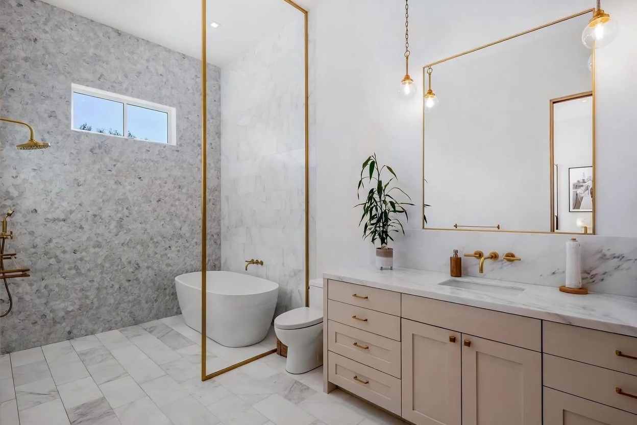 This modern primary bathroom features a sleek, open-concept wet room area with a freestanding tub enclosed by a gold-framed glass partition. The space is finished with light-toned stone surfaces, a double-sink vanity with gold hardware, and a large mirror illuminated by hanging pendant lights. The overall aesthetic is bright, luxurious, and contemporary, emphasizing clean lines and high-end finishes.