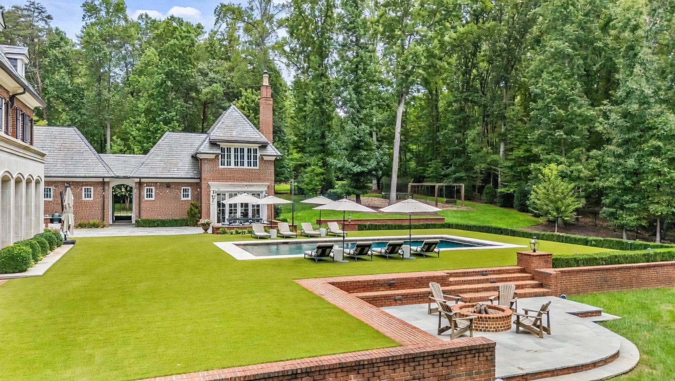 This expansive rear view of a luxury brick estate showcases a meticulously landscaped backyard featuring a rectangular swimming pool surrounded by lounge chairs and umbrellas. A sunken stone patio with a circular fire pit provides an intimate seating area, while the lush green lawn transitions into a dense, mature forest backdrop. The architectural design includes a prominent brick chimney and classic multi-pane windows, creating a sophisticated and serene outdoor living environment.
