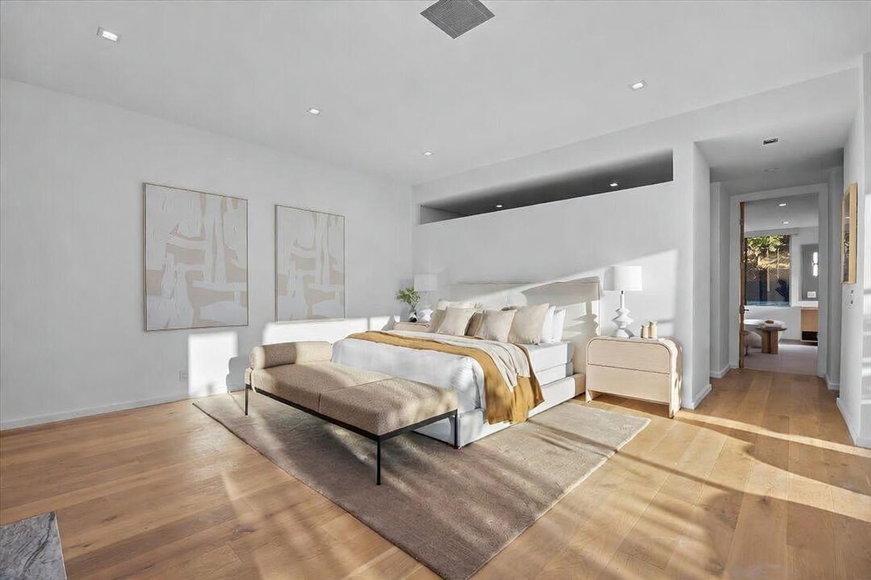 This spacious primary bedroom features light wood flooring, neutral walls, and a large bed centered in the room with a bench at the foot. Two abstract art pieces hang on the left wall, while a doorway leads to an adjacent room, creating an open and airy atmosphere. The room is illuminated by recessed lighting and soft natural light, contributing to a serene and sophisticated aesthetic.