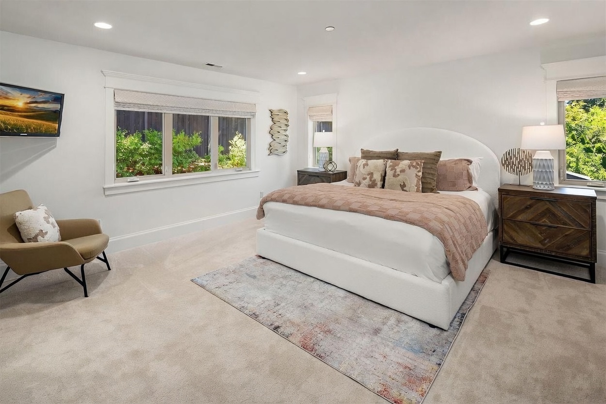 This spacious primary bedroom features a neutral color palette with soft carpeting and white walls, creating a serene and inviting atmosphere. A large, upholstered bed serves as the focal point, complemented by a patterned area rug, a modern armchair, and two wooden nightstands with matching lamps. Large windows allow for ample natural light and provide a view of the lush greenery outside.