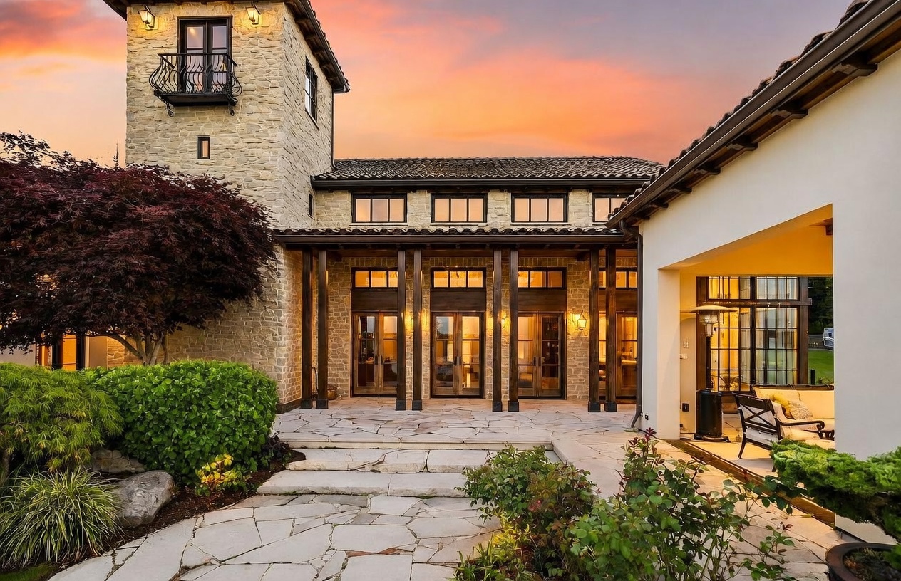 This elegant Mediterranean-style home features a stone-clad exterior with a prominent tower and a covered patio area. The scene is captured at sunset, highlighting the warm glow from the interior lighting reflecting off the large glass doors and windows. A flagstone patio leads to the home, framed by lush landscaping and a mature tree, creating a sophisticated and inviting outdoor living space.