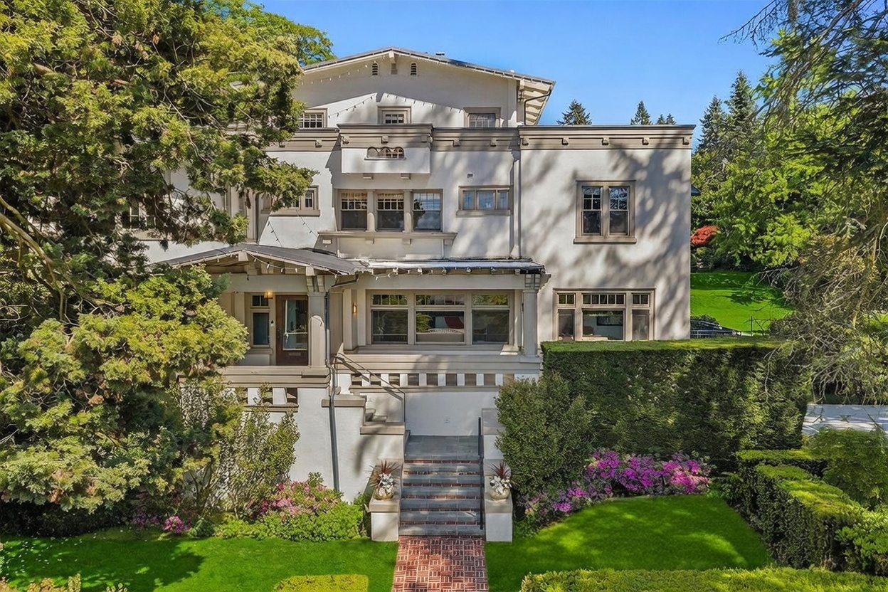 This stately, multi-story residence features a classic architectural design with a light-colored stucco facade and symmetrical window placement. A prominent covered porch with a brick-paved walkway leads to the main entrance, while a set of stone steps provides access from the lush, manicured lawn. The property is surrounded by mature trees and well-maintained hedges, creating a private and elegant curb appeal.