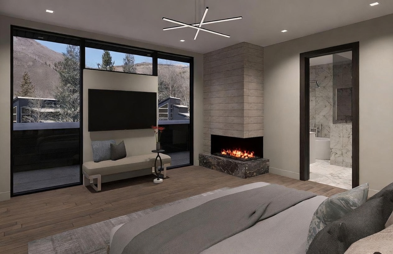 This modern primary bedroom features a sleek, minimalist aesthetic with light wood flooring and a neutral color palette. A corner fireplace with a dark stone hearth and a tall, textured stone surround serves as a focal point, while large windows provide expansive views of the snowy mountain landscape. The room includes a contemporary bench, a wall-mounted television, and an open doorway leading into a bright, marble-tiled bathroom.