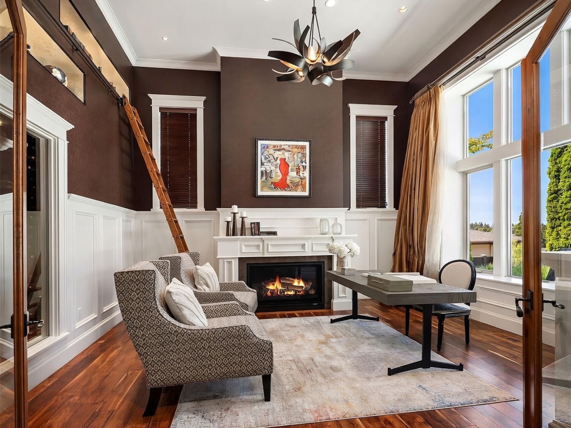 This sophisticated home office features dark brown walls contrasted by crisp white wainscoting and a matching fireplace mantel. A rolling library ladder leans against the wall, leading to an upper storage shelf, while a modern desk and two patterned armchairs create a functional workspace. Large windows on the right side of the room flood the space with natural light, highlighting the rich wood flooring and the contemporary chandelier above.