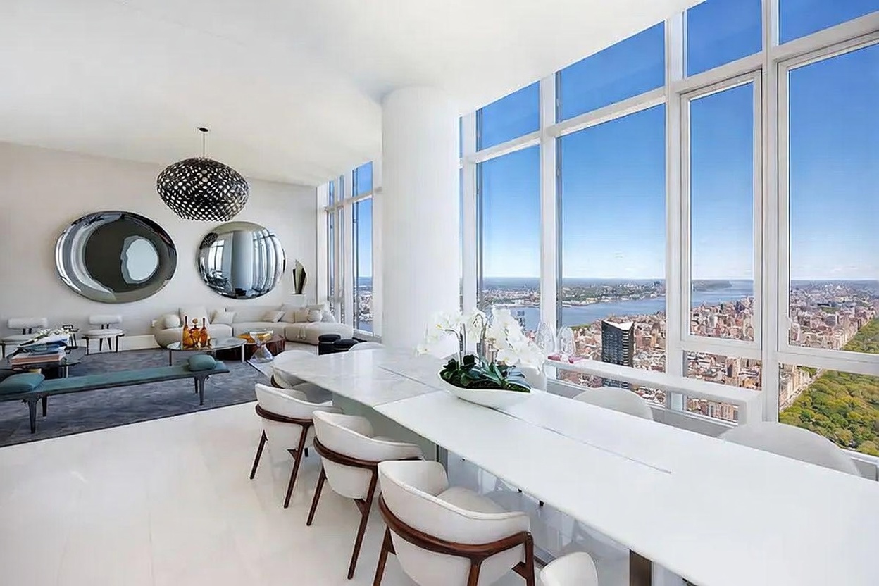 This expansive, high-rise dining and living area features a sleek, long white dining table paired with modern chairs, positioned alongside floor-to-ceiling windows that offer a panoramic view of the city skyline and river. The adjacent living space is furnished with a neutral-toned sectional sofa, a low-profile bench, and two large circular wall mirrors, all illuminated by a striking, dark geometric pendant light. The room is characterized by its bright, minimalist aesthetic, polished light-colored flooring, and an open-concept layout that emphasizes the dramatic urban vistas.