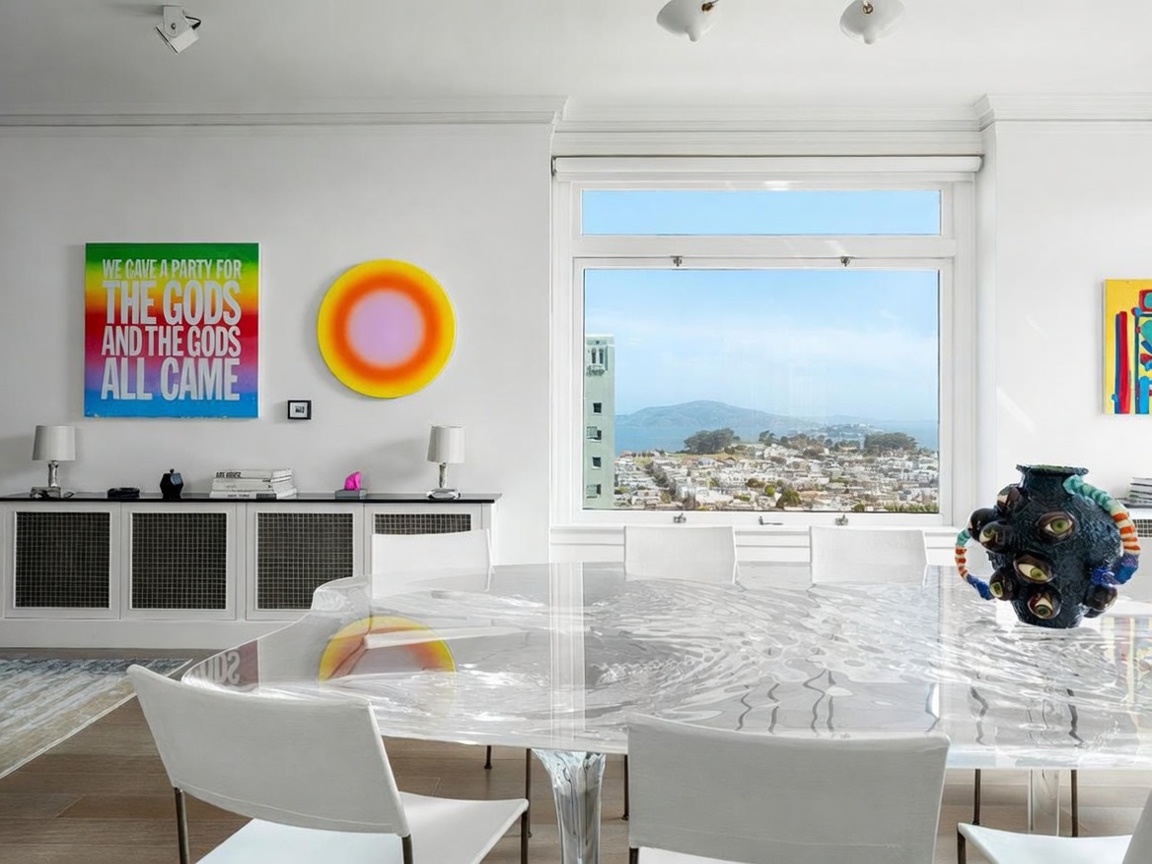This bright and modern dining room features a unique, transparent, water-textured table as the centerpiece, surrounded by minimalist white chairs. The room is adorned with vibrant, contemporary art pieces on the walls and a sleek white sideboard, while a large window provides a stunning, elevated view of the city and bay. The overall aesthetic is sophisticated and artistic, emphasizing clean lines and a bright, airy atmosphere.