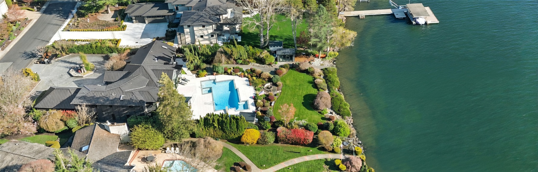 This high-angle aerial view captures a luxurious waterfront estate, showcasing a sprawling main residence with a dark metal roof, a private swimming pool, and meticulously landscaped grounds. The property features a long private dock extending into the calm lake, surrounded by lush greenery and mature trees. The perspective highlights the seamless integration of high-end residential architecture with the natural beauty of the shoreline.