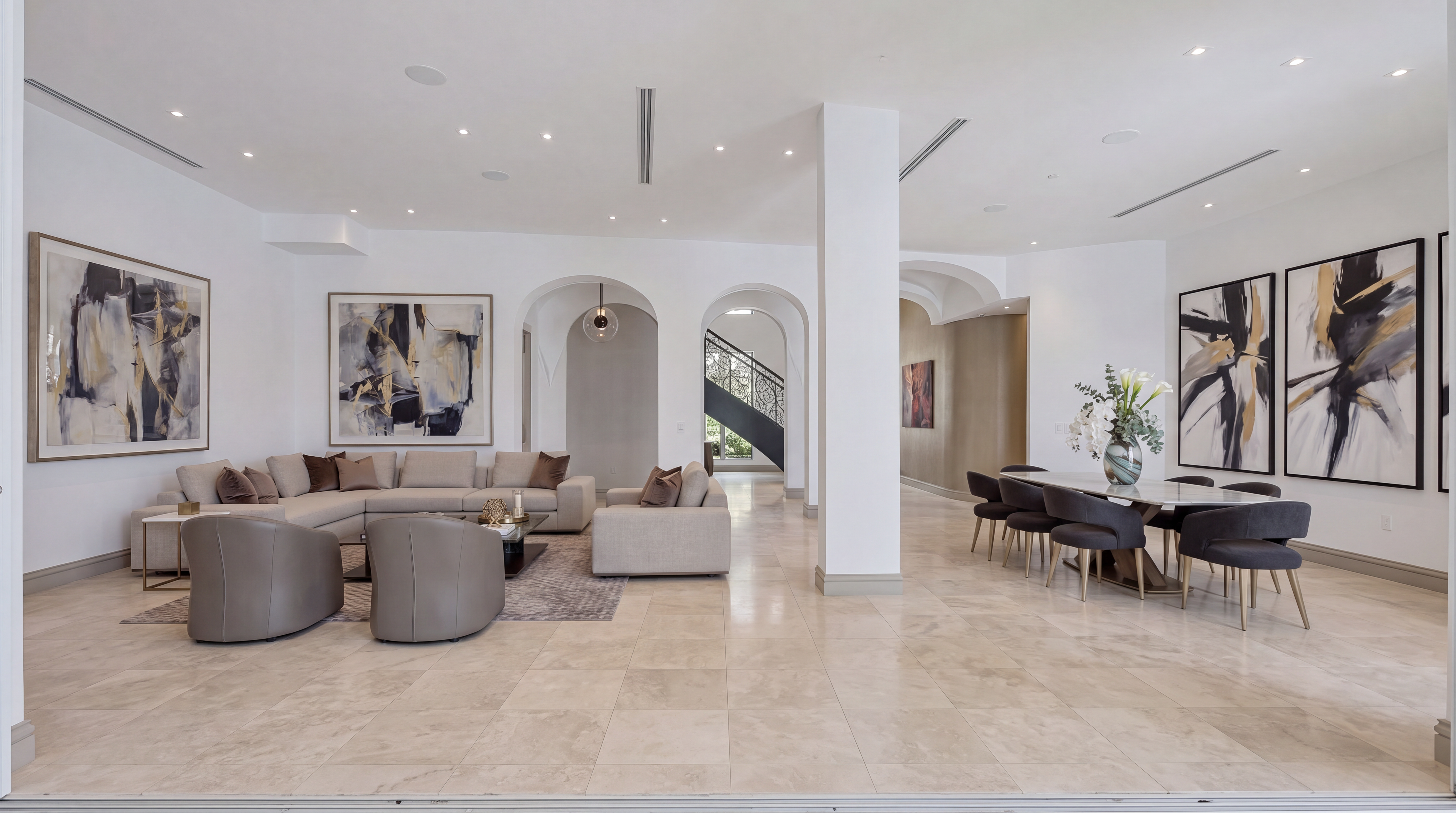 This spacious, open-concept living and dining area features a bright, sophisticated aesthetic with neutral tones and elegant tile flooring. The space is highlighted by a large sectional sofa, contemporary leather armchairs, and formal dining seating, all complemented by bold abstract artwork. Arched architectural details and clean lines create a seamless flow, making it ideal for both relaxed living and upscale entertaining.