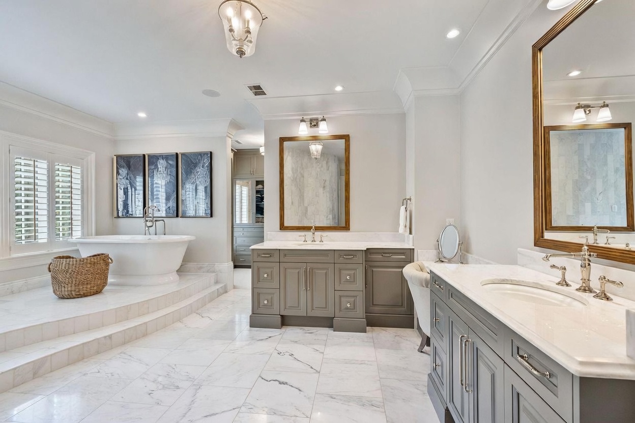 This luxurious primary bathroom features a spacious layout with dual vanities, a freestanding soaking tub set on a raised platform, and elegant stone flooring. The room is accented by gold-toned fixtures, framed mirrors, and a neutral color palette that creates a sophisticated and serene atmosphere. The perspective captures the entire vanity area and the elevated tub nook, highlighting the high-end finishes and open design.