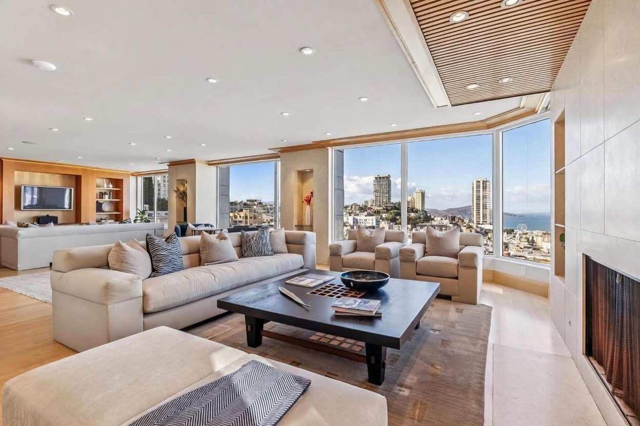 This expansive, light-filled living room features a neutral color palette with plush sofas and armchairs arranged around a large, dark wood coffee table. The space is defined by its impressive wall of windows that offer panoramic city and bay views, complemented by a unique slatted wood ceiling detail. A stone-clad chimney breast anchors the room, creating a sophisticated and airy atmosphere perfect for entertaining.
