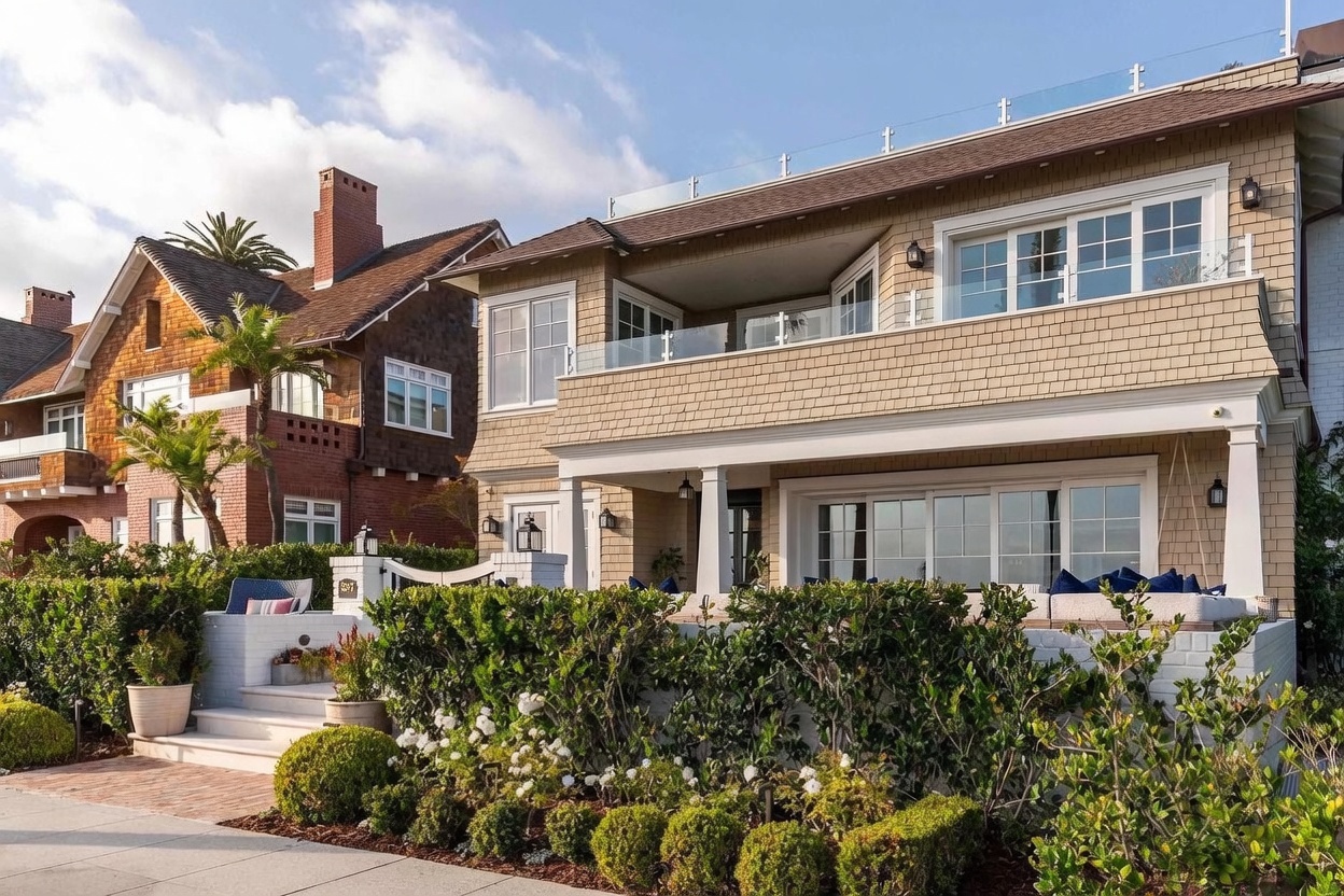 This charming coastal-style home features a prominent front porch with white columns and a spacious second-story balcony enclosed by glass railings. The exterior is finished in light-toned shingles, complemented by a neatly manicured hedge and a brick-paved walkway leading to the entrance. The property exudes a welcoming, upscale beach-house aesthetic, with a neighboring historic brick home visible to the left.