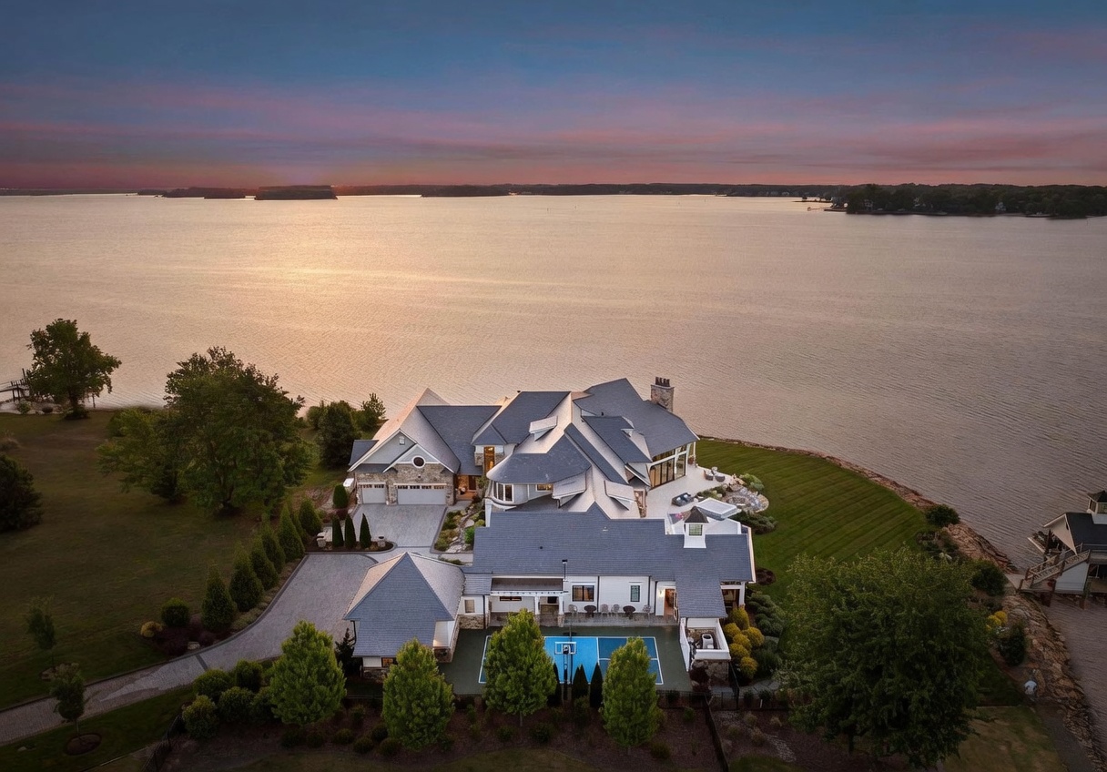 This stunning aerial view captures a sprawling luxury estate situated directly on a expansive lakefront property at sunset. The multi-gabled residence features a complex roofline, a paved motor court, and a private sports court, all surrounded by manicured lawns and mature trees. The composition highlights the seamless integration of the home with its natural waterfront setting, emphasizing the expansive views and serene atmosphere.