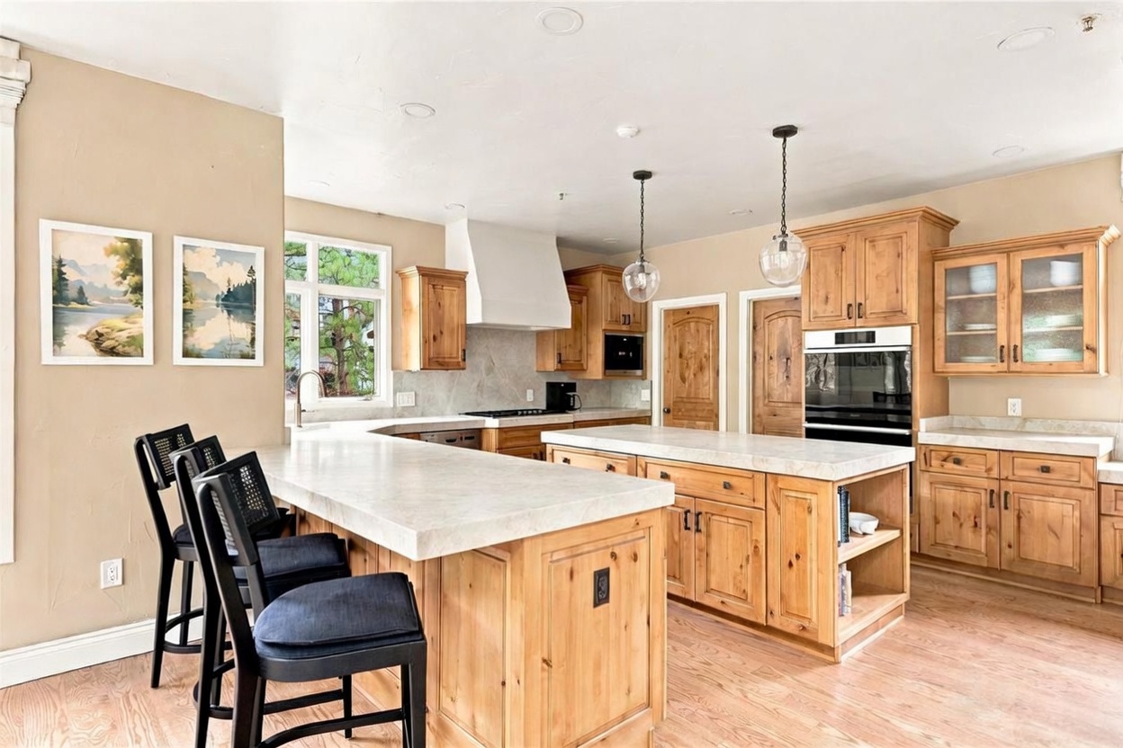 This spacious kitchen features light wood cabinetry, a large central island with a stone countertop, and a breakfast bar with three dark-toned stools. The space is illuminated by two glass pendant lights and includes stainless steel appliances, a white range hood, and a window overlooking the outdoors. The warm wood flooring and neutral wall tones create a welcoming and functional culinary environment.