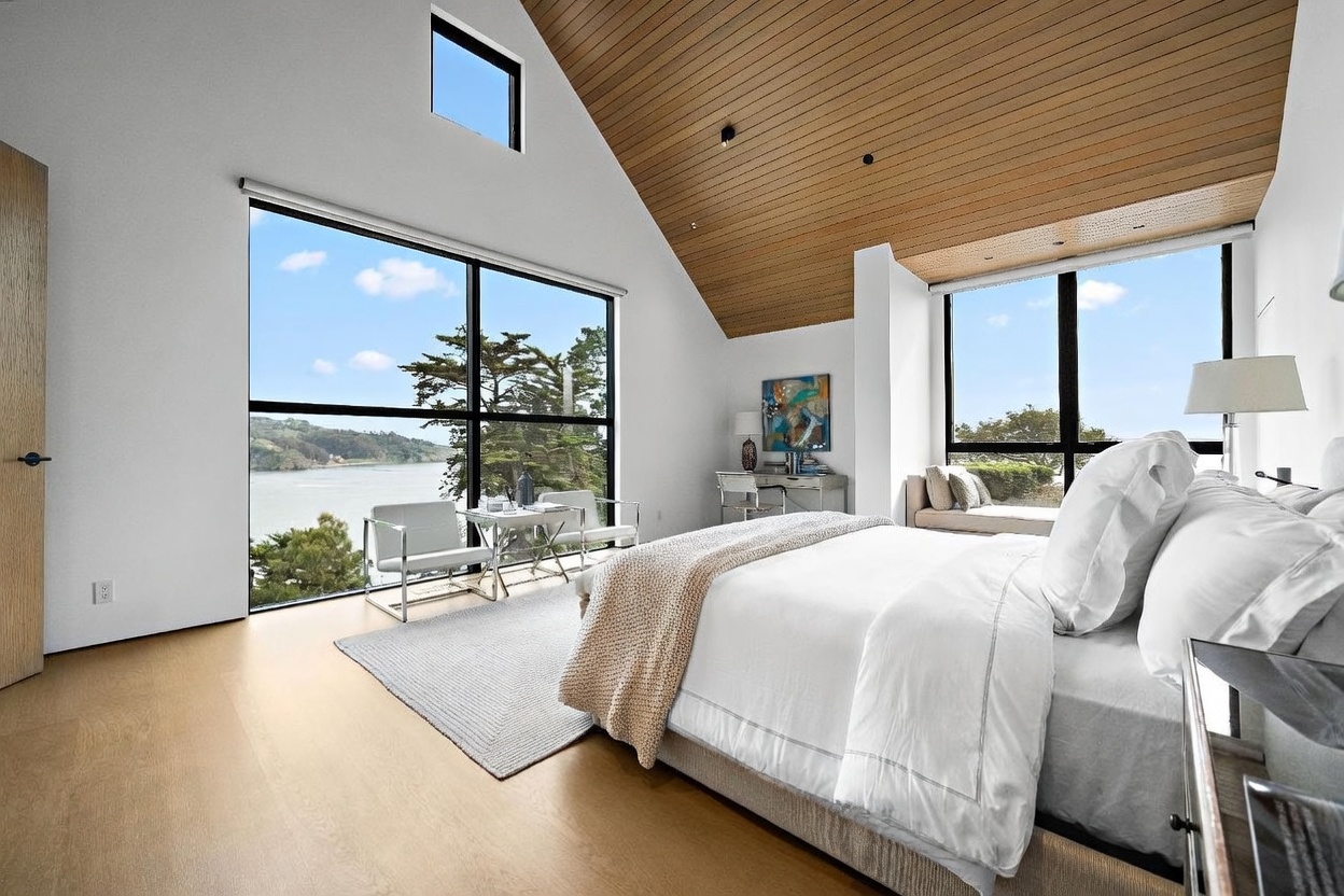 This bright and airy primary bedroom features a vaulted ceiling with light wood paneling and large windows that offer expansive water views. The room is furnished with a plush bed, a cozy window seat, and a small desk area, all set against a clean, minimalist white backdrop. The perspective captures the serene atmosphere and the seamless integration of the natural landscape into the living space.