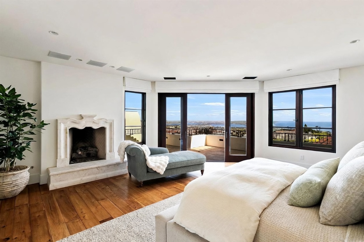This primary bedroom features a light-filled, airy aesthetic with hardwood flooring and a neutral color palette. A stone fireplace with a decorative mantel sits to the left, while a blue chaise lounge is positioned near large glass doors that open onto a private balcony with expansive ocean views. The room is anchored by a large bed with textured bedding, creating a serene and luxurious atmosphere.