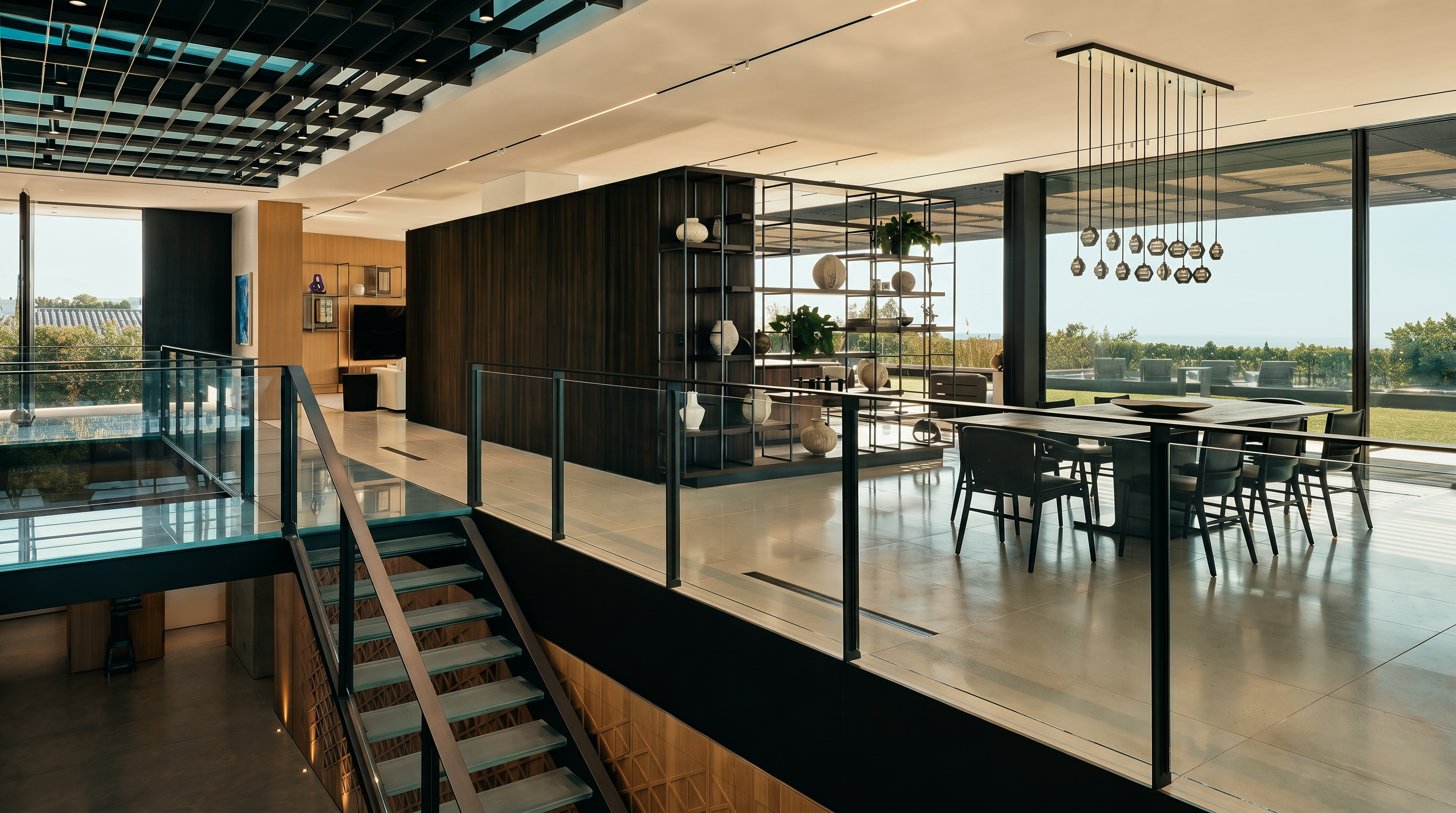 This sophisticated, open-concept dining area features a sleek wooden table surrounded by modern black chairs, illuminated by a striking linear pendant light fixture. The space is defined by a glass-railed staircase and a dark wood partition wall with integrated shelving, creating a seamless transition between levels. Large floor-to-ceiling windows offer an expansive view of the outdoor landscape, emphasizing the home's contemporary architectural design and airy, light-filled atmosphere.