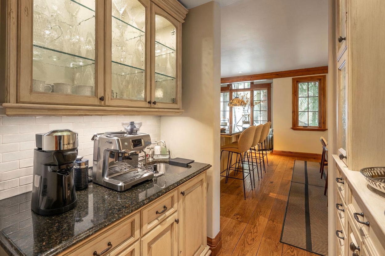 This image captures a well-appointed coffee bar area featuring light wood cabinetry, dark stone countertops, and a white subway tile backsplash. A professional-grade espresso machine and a pod coffee maker sit on the counter, adjacent to a small integrated sink. The perspective looks through a transition space into a dining area with bar seating, warm wood flooring, and large windows that bring in natural light.