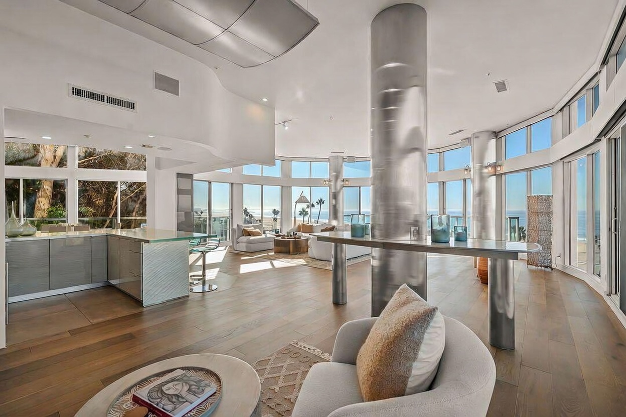 This expansive, open-concept living area features a modern aesthetic with light wood flooring and a prominent metallic structural column. The space is defined by large windows that provide panoramic ocean views, seamlessly connecting the interior to the coastal environment. A contemporary armchair sits in the foreground, while the background showcases a bright, airy living room layout with a kitchen island visible to the left.
