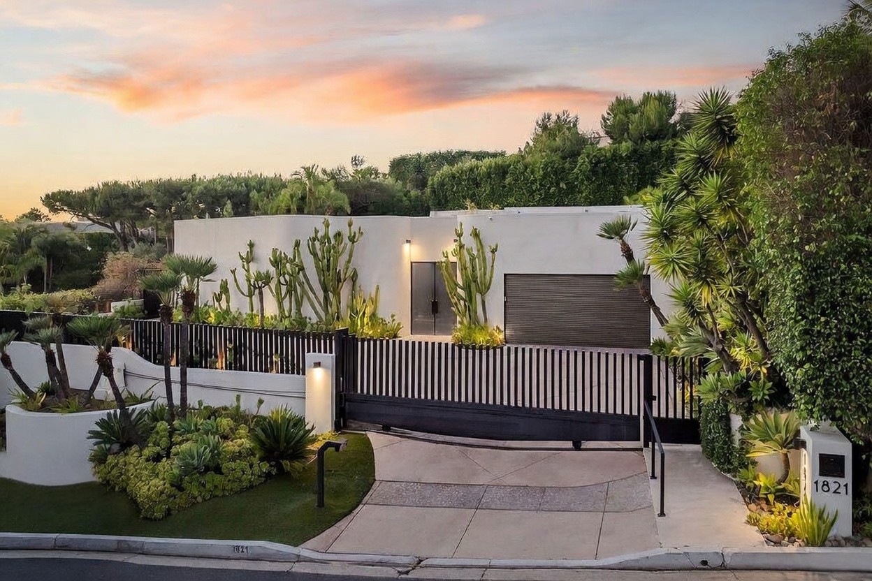 This modern, minimalist residence features a smooth, white-stucco facade accented by a variety of desert-inspired landscaping, including tall cacti and lush greenery. A sleek, dark-toned horizontal slat gate provides privacy for the driveway, while the property is framed by mature trees under a vibrant sunset sky. The perspective is a ground-level shot looking toward the gated entrance, highlighting the home's contemporary curb appeal and architectural simplicity.
