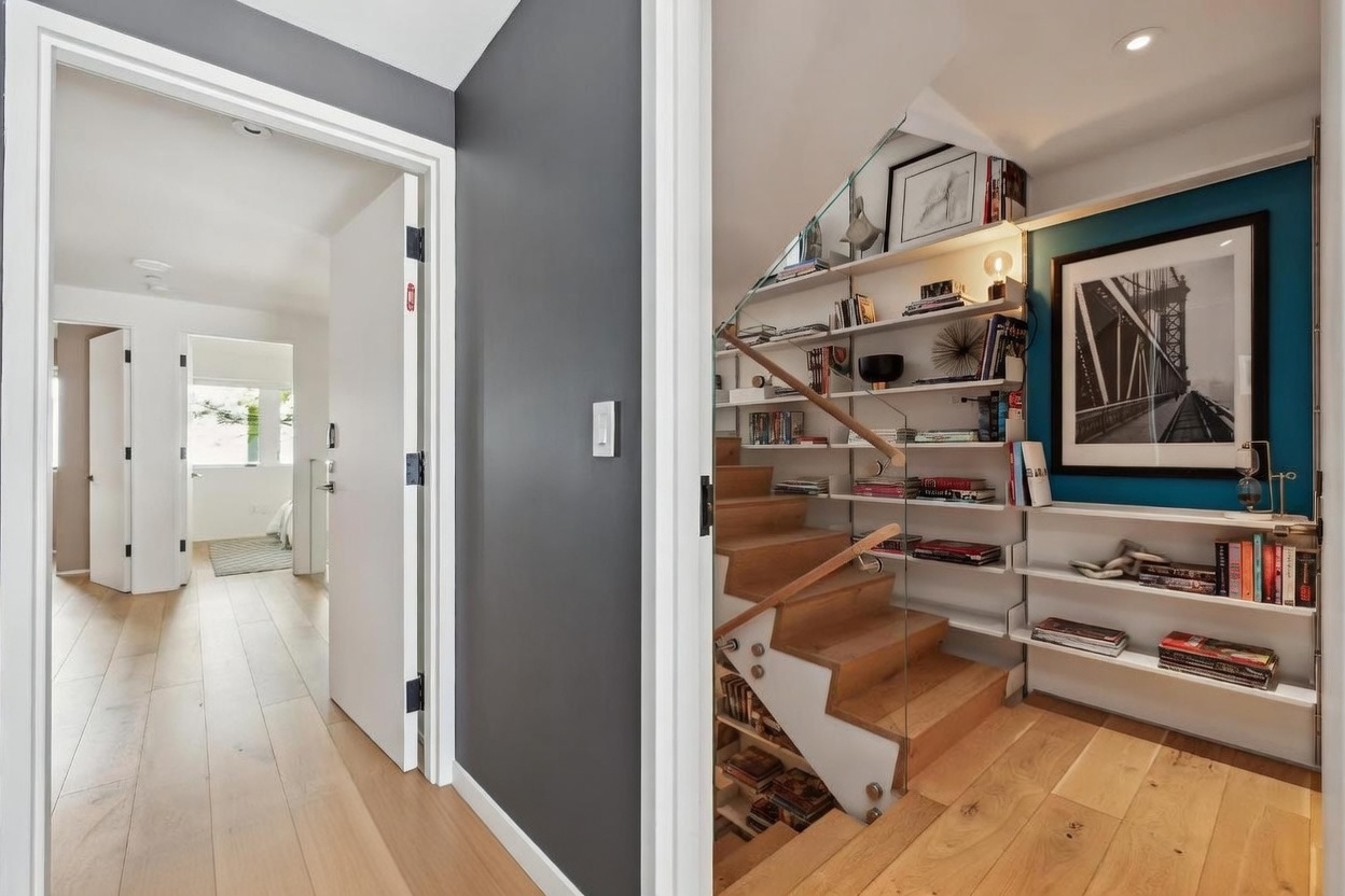 This interior view captures a transition space featuring a staircase with light wood treads and a glass railing, adjacent to a built-in shelving unit that displays books and decorative items. To the left, a doorway leads into a bright bedroom with light wood flooring, while the foreground shows a tiled entryway floor. The space is characterized by a modern, clean aesthetic with a bold dark accent wall and a blue-painted wall behind the shelving.