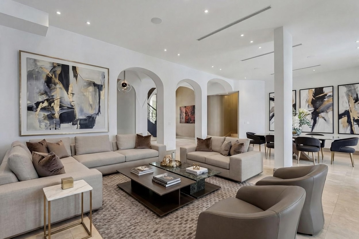 This sophisticated living room features a spacious open-concept layout with neutral-toned sofas and modern leather armchairs arranged around a tiered coffee table. The space is defined by elegant arched architectural openings that lead to other areas of the home, complemented by large abstract artwork on the walls. The room is bathed in soft recessed lighting, creating a bright and airy atmosphere that flows seamlessly into an adjacent dining area.