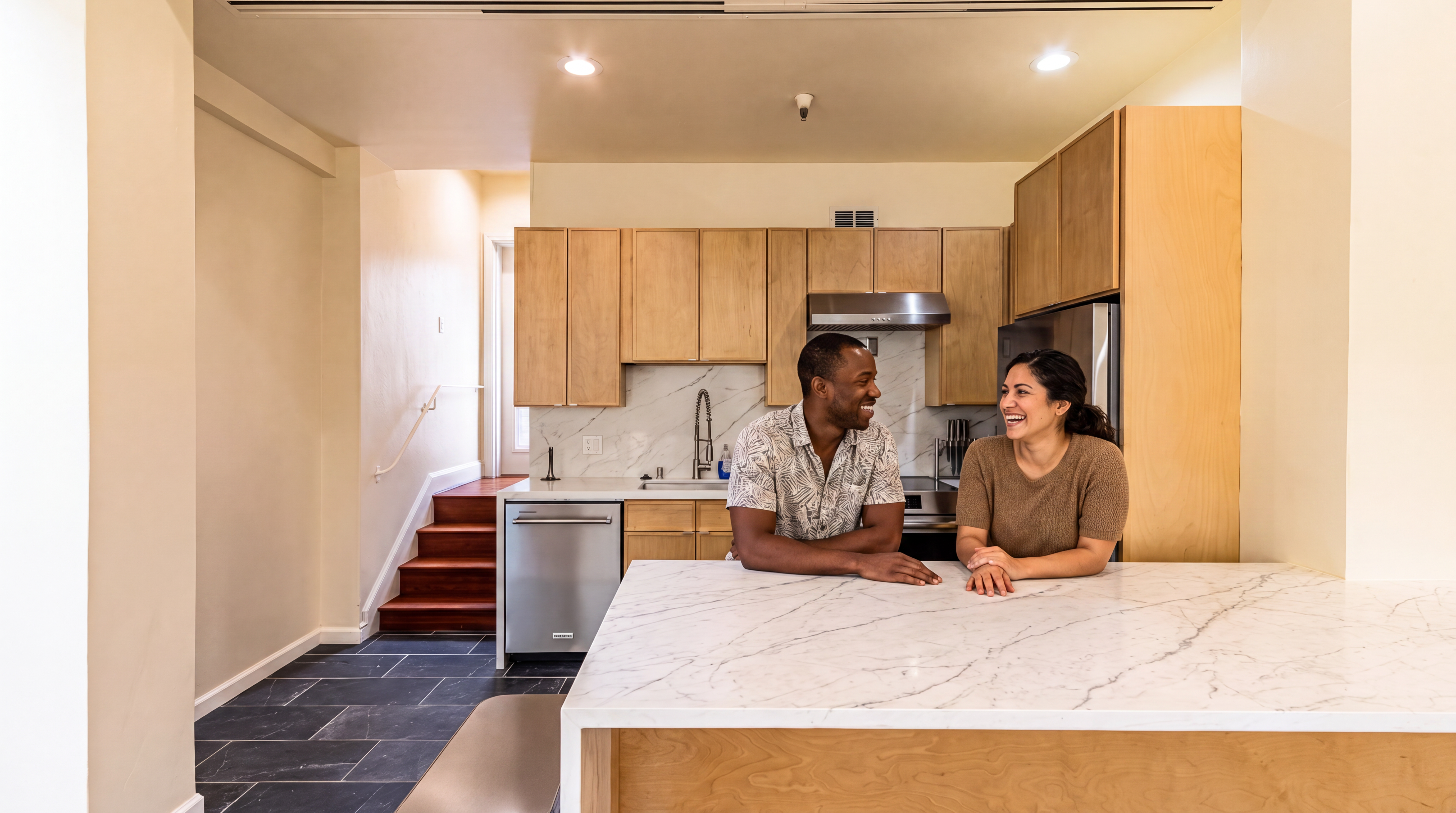This bright, contemporary kitchen features natural wood cabinetry, sleek marble countertops, and a full suite of stainless steel appliances. The open-concept design seamlessly integrates with a stone-tiled floor and a wooden staircase, creating a warm, inviting atmosphere. A couple is shown gathered around the large, marble-topped center island, highlighting the kitchen's functionality as a social hub for the home.