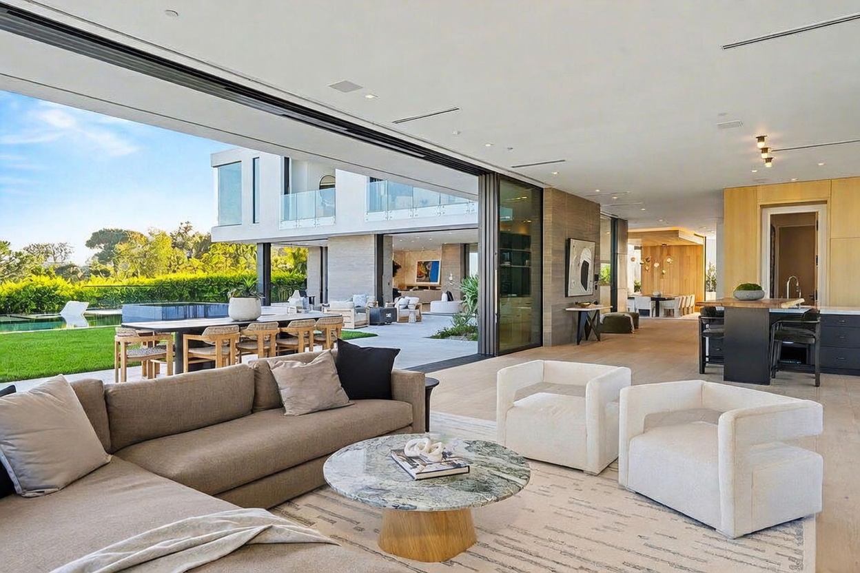 This expansive, open-concept living area features a seamless transition to the outdoors through large, retractable glass walls. The space is furnished with a neutral-toned sectional sofa, two modern armchairs, and a circular stone-topped coffee table, all set upon a textured area rug. The interior design emphasizes a bright, airy atmosphere with light wood flooring and a view that extends past the patio to a private pool and lush landscaping.
