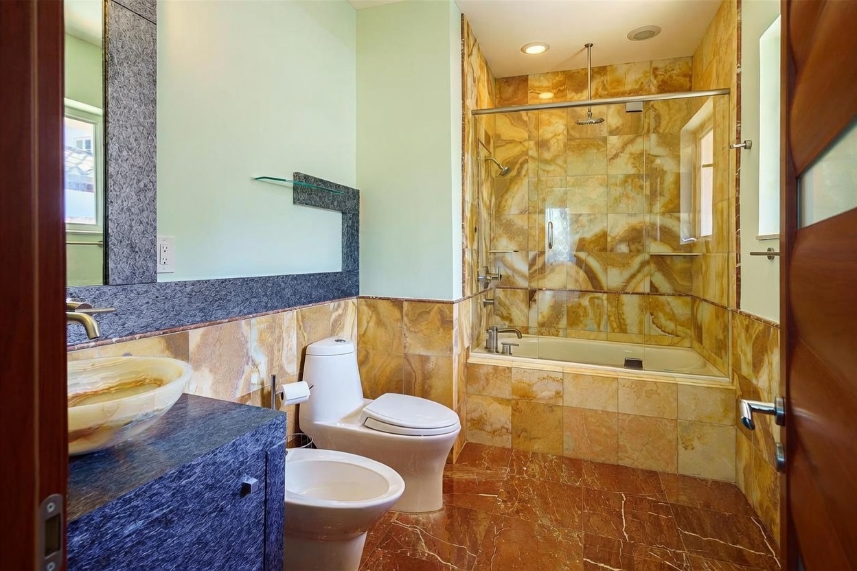 This bathroom features a combination tub and shower enclosure with warm-toned, veined stone tile walls and a glass door. A pedestal toilet and a bidet are positioned next to a vanity with a dark, textured stone countertop and a vessel sink. The room is finished with matching stone floor tiles and light-colored walls, creating a cohesive and warm aesthetic.