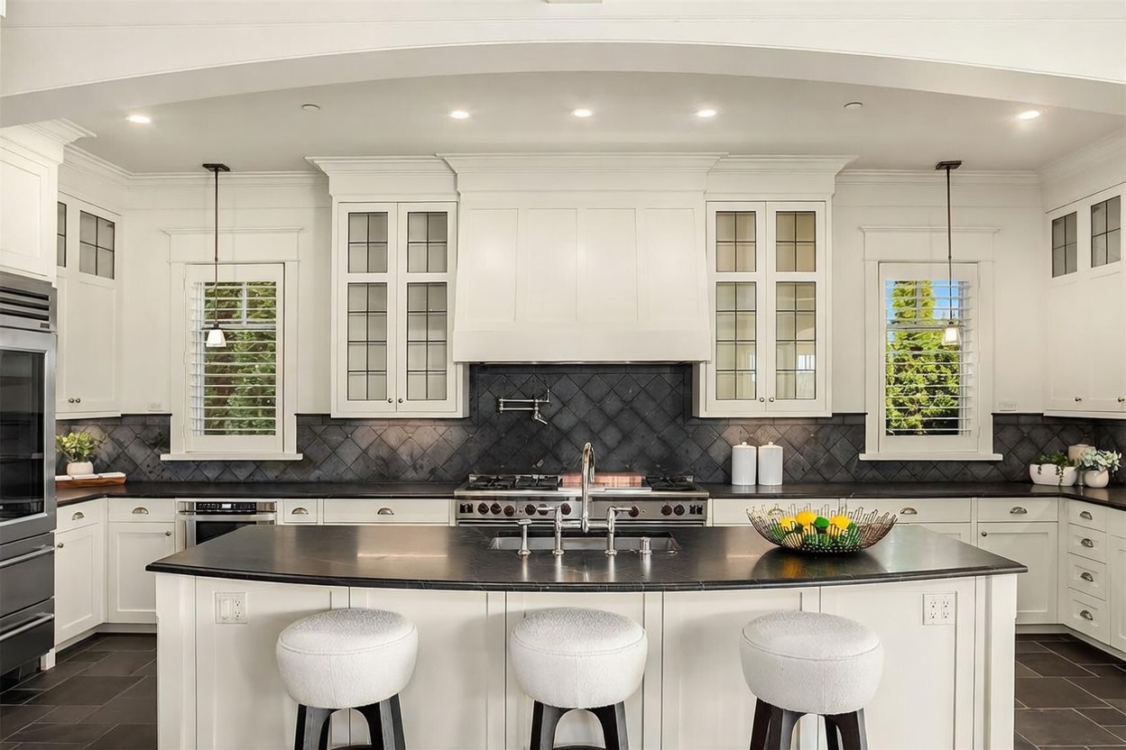 This elegant, high-end kitchen features crisp white cabinetry, a dark stone-topped island with three upholstered stools, and a striking dark-tiled backsplash. The symmetrical layout is anchored by a professional-grade range and a custom hood, while pendant lighting and plantation shutters add a refined, classic touch. The perspective is a centered, eye-level shot that emphasizes the spaciousness and sophisticated design of the culinary space.