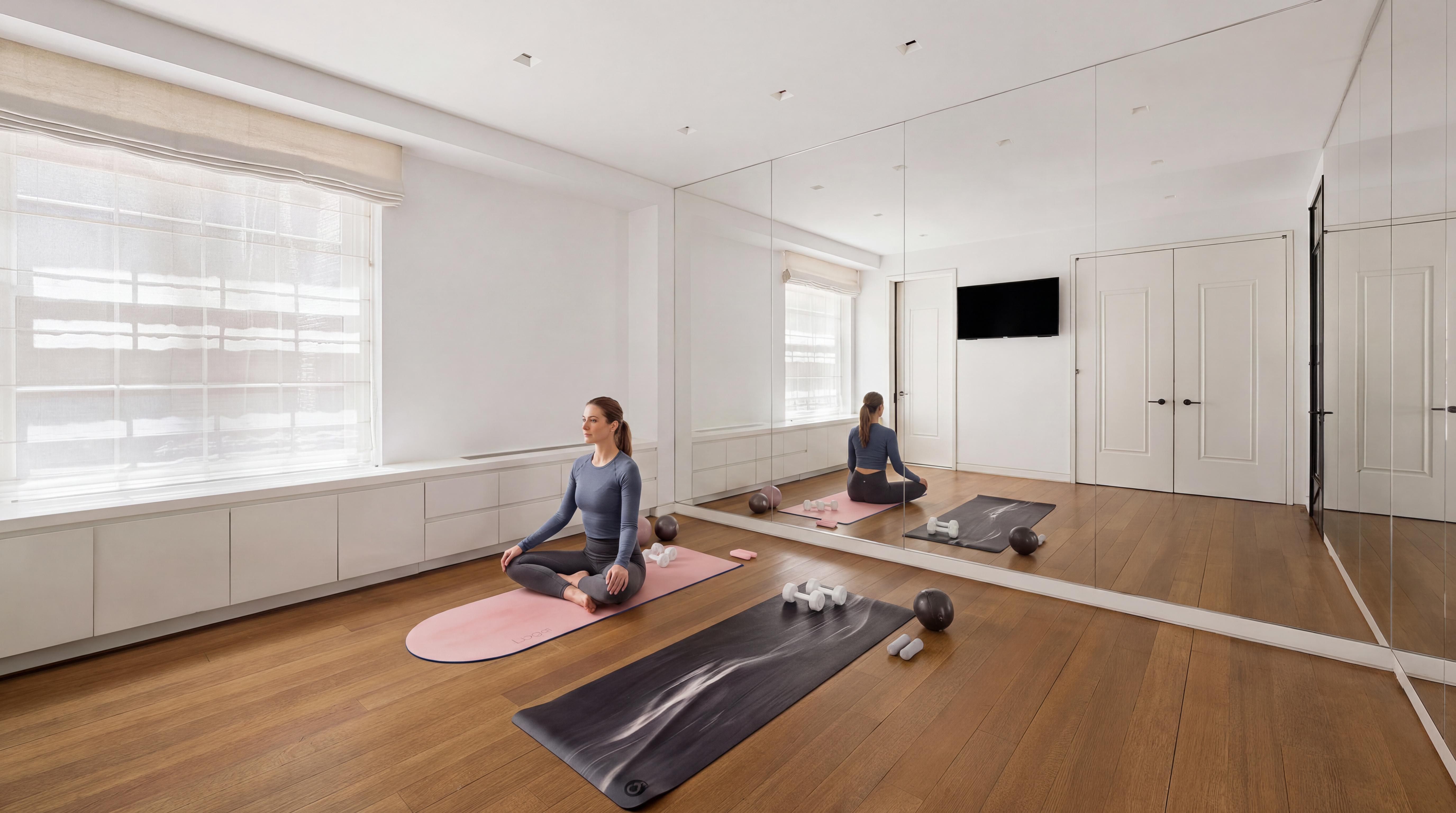 This sleek and minimalist home gym features warm hardwood flooring, a large wall-to-wall mirror that enhances the sense of space, and expansive windows fitted with clean Roman shades. The room includes two yoga mats, light dumbbells, and balance balls, creating an inviting atmosphere for fitness and wellness activities. The crisp white color palette and thoughtful lighting contribute to a bright, serene, and professional aesthetic.