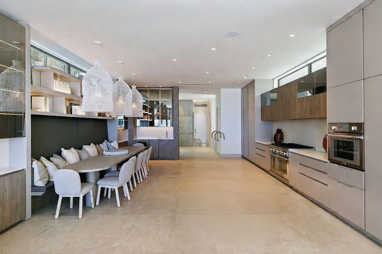 This contemporary open-concept kitchen and dining area features a long central island with a light-toned wood base and a smooth countertop. To the left, a built-in banquette seating arrangement with a long table is illuminated by three large, textured pendant lights. The space is finished with neutral-toned cabinetry, integrated appliances, and large-format floor tiles, creating a cohesive and sophisticated modern aesthetic.