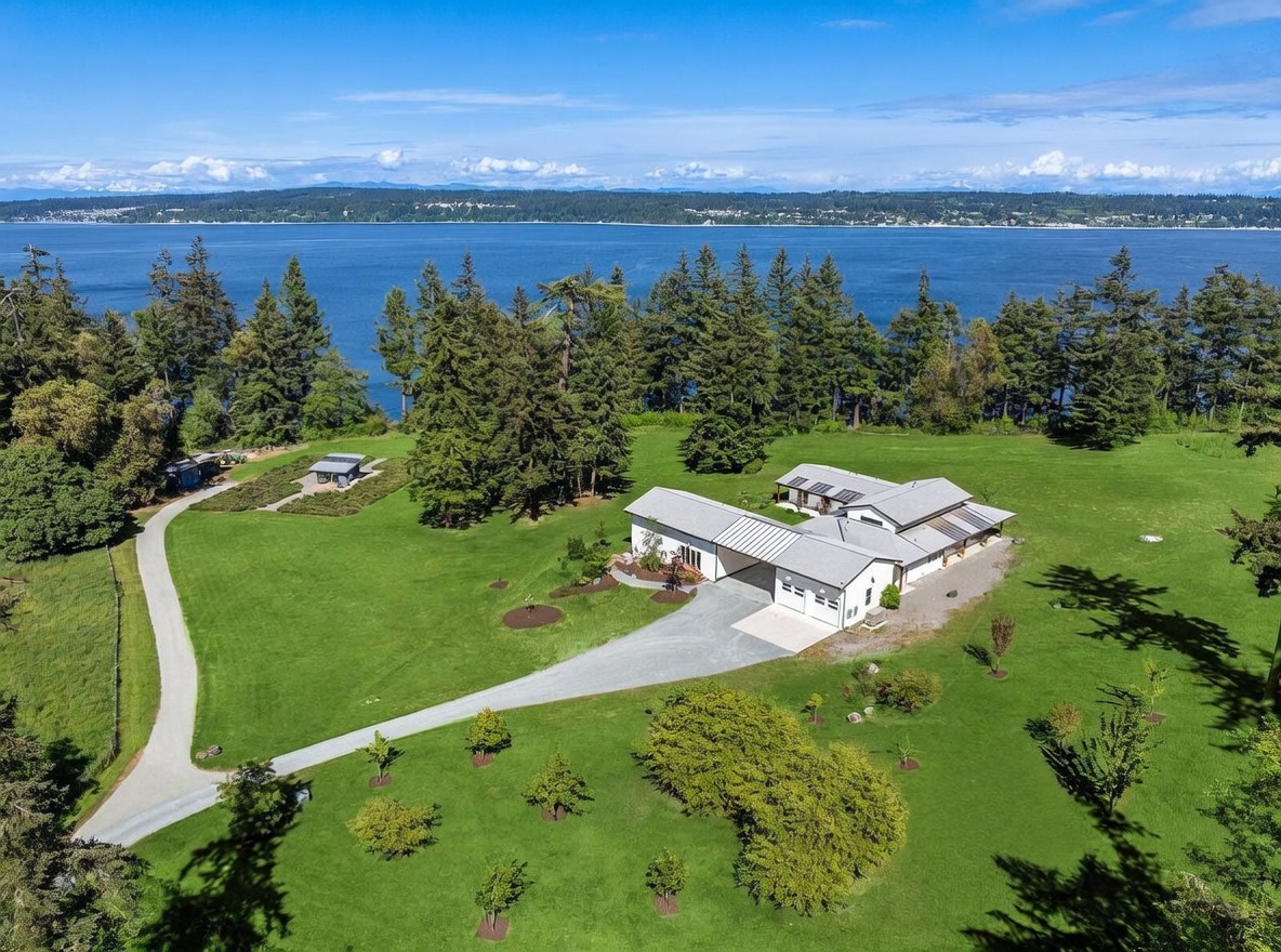 This aerial view captures a sprawling, modern farmhouse-style residence situated on a large, manicured grassy lot overlooking a vast body of water. The property features a long, winding gravel driveway, a detached outbuilding, and a fenced garden area, all surrounded by dense evergreen trees. The home itself boasts a multi-gabled roofline, a spacious attached garage, and expansive outdoor living spaces that take full advantage of the scenic waterfront location.