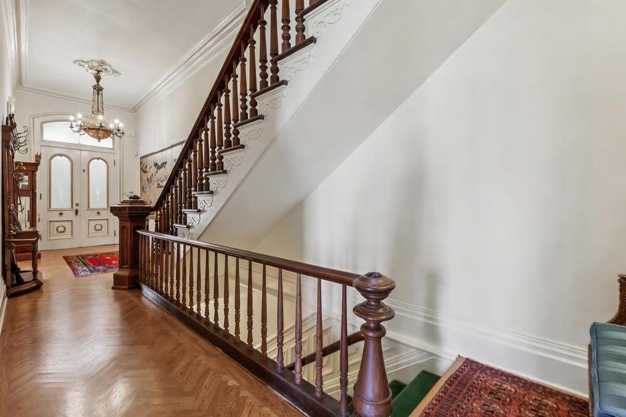 This elegant entryway features a grand staircase with intricate dark wood railings and balusters, set against a backdrop of crisp white walls. The herringbone-patterned wood flooring leads toward a classic double-door entrance illuminated by a decorative chandelier. A rich, patterned rug adds a touch of warmth to the space, creating a sophisticated and welcoming first impression.