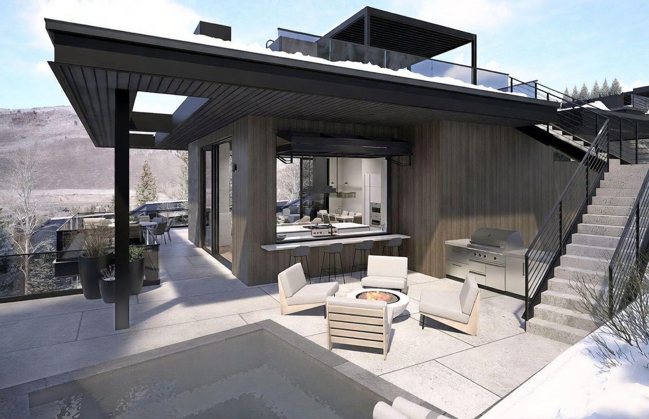 This expansive, modern outdoor terrace features a sleek, covered lounge area complete with a central fire pit and comfortable seating. The space seamlessly integrates with an indoor kitchen visible through a large pass-through window, while a stainless steel grill and concrete stairs leading to an upper level enhance the functional design. The setting offers a sophisticated, contemporary aesthetic with panoramic views of the surrounding mountainous landscape.