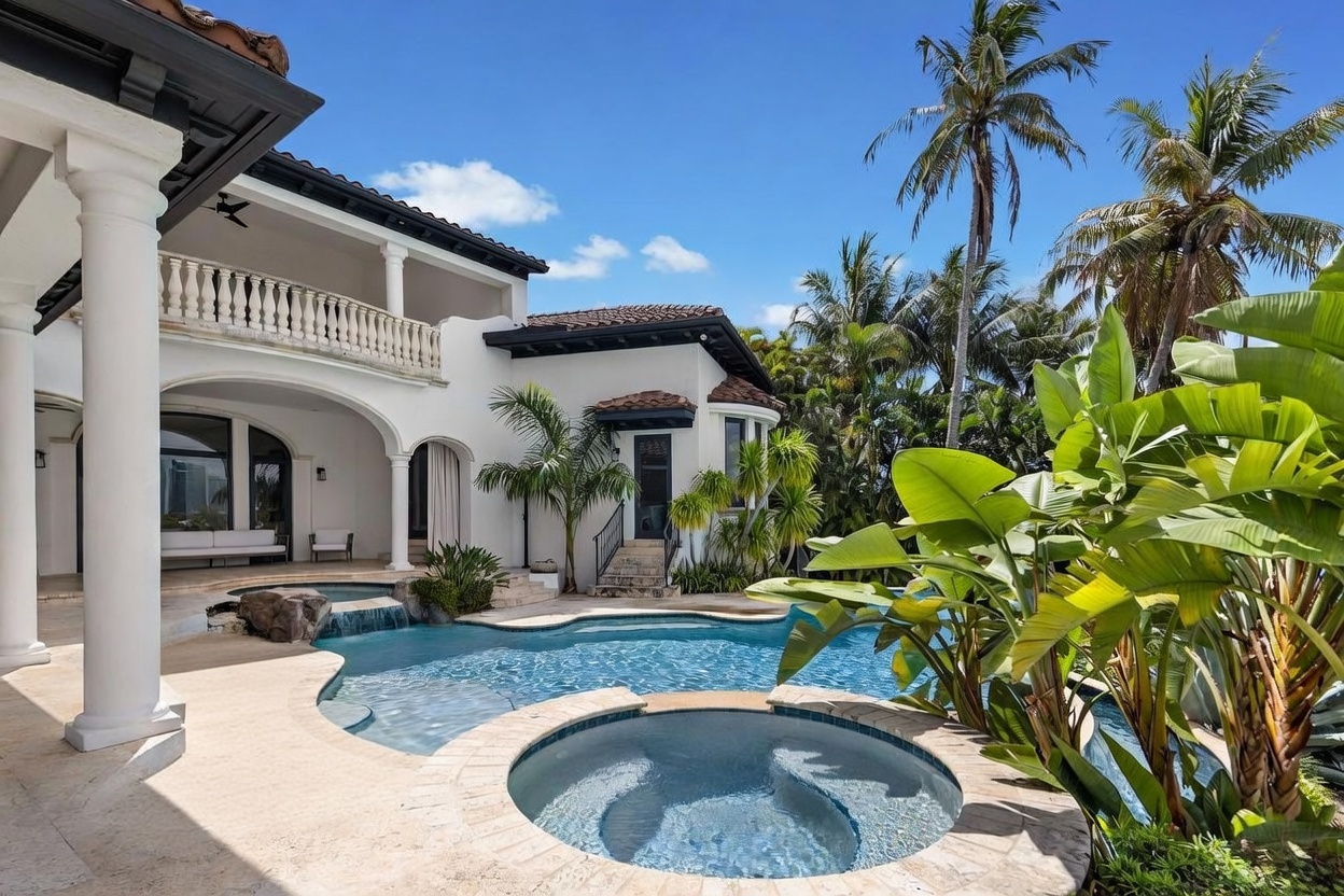This luxurious outdoor living space features a custom-shaped swimming pool with a built-in spa and a cascading rock waterfall feature. The white stucco home provides a Mediterranean-inspired backdrop, complete with a covered patio area and a second-story balcony with a stone balustrade. Lush tropical landscaping, including tall palm trees and vibrant green foliage, frames the scene, creating a serene and private resort-like atmosphere.