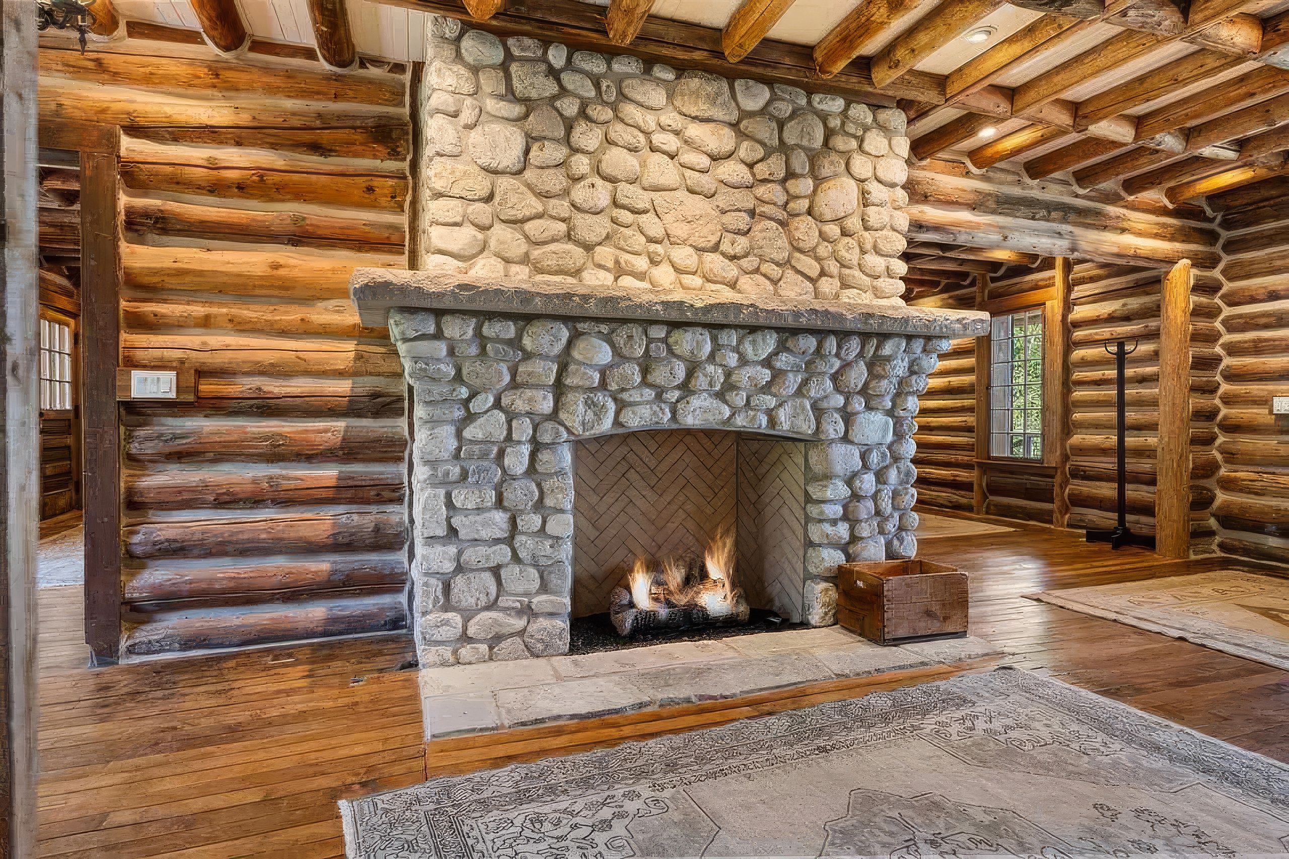 This rustic living room features a prominent stone-clad chimney breast with a substantial stone mantel and a herringbone-patterned firebox. The space is defined by its authentic log walls, exposed timber ceiling beams, and warm wood flooring accented by a large patterned area rug. The perspective captures the hearth as the central focal point, emphasizing the cozy, cabin-inspired aesthetic of the home.