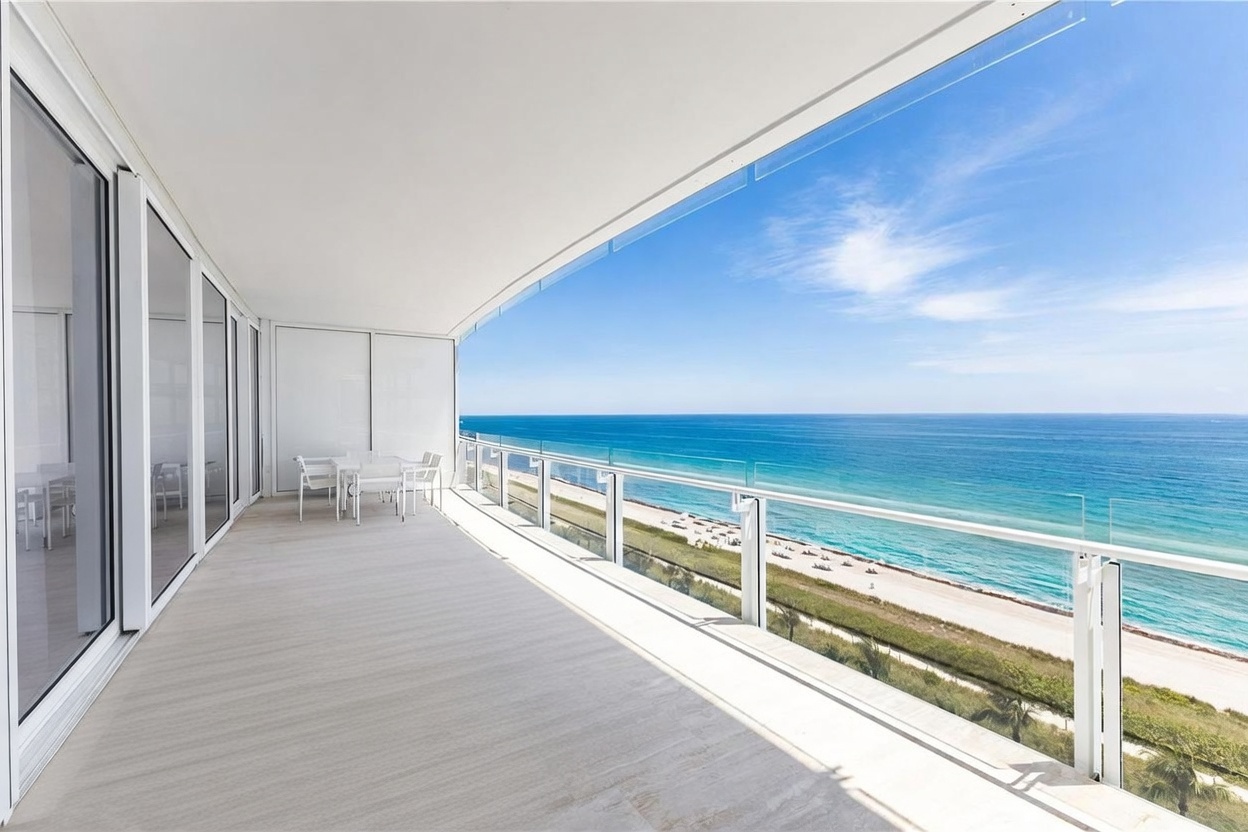 This expansive, modern balcony offers a panoramic view of the ocean and sandy beach, framed by a sleek glass railing. The space is furnished with white outdoor seating, including a table and chairs, creating a serene area for relaxation. The clean, minimalist design emphasizes the seamless transition between the indoor living area and the coastal scenery.