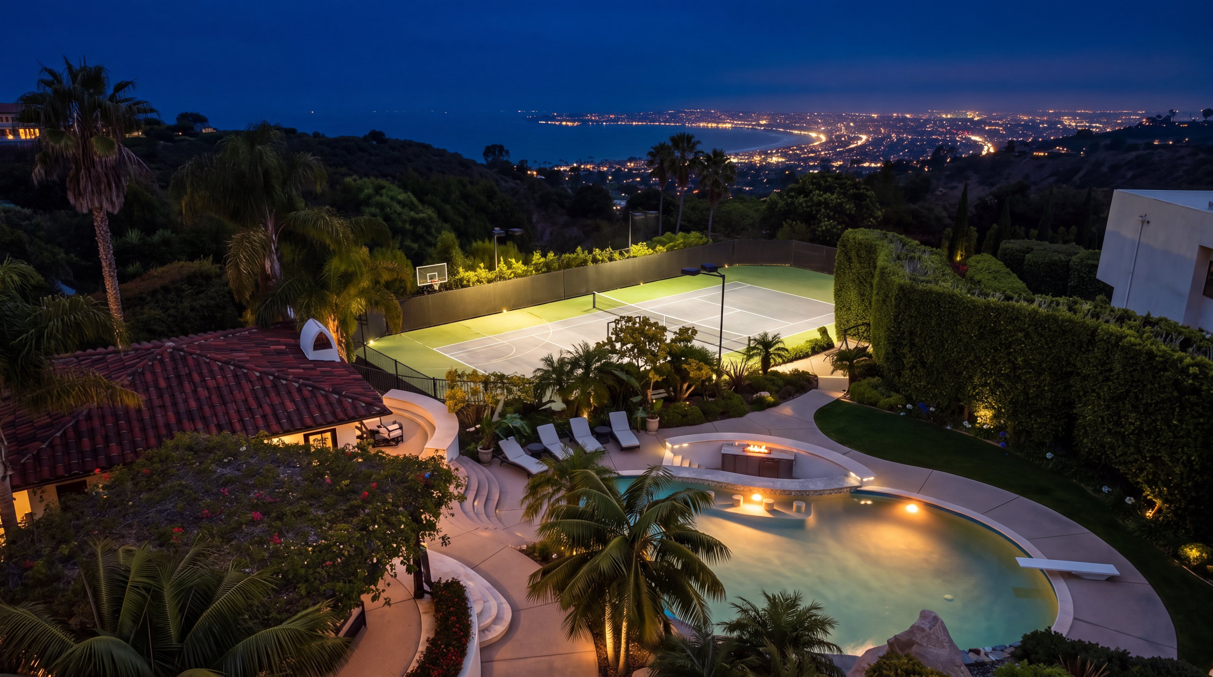 This breathtaking aerial night view captures a luxurious estate featuring a meticulously landscaped backyard complete with an illuminated swimming pool, built-in fire pit, and a private tennis court. The property offers stunning panoramic views of the distant coastal city lights against a dark evening sky. The integration of high-end outdoor living spaces and recreational facilities creates an exclusive and tranquil atmosphere perfect for entertaining.
