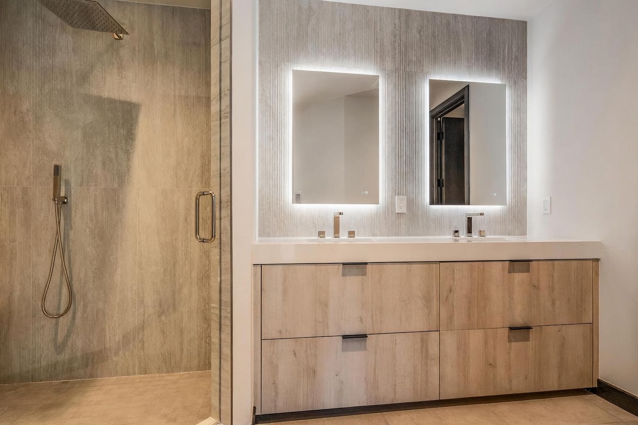 This is a primary bathroom featuring a double vanity with light wood cabinetry and a white countertop. Two illuminated mirrors hang above the sinks, set against a textured gray wall. A glass-enclosed shower is visible to the left, showcasing modern fixtures and tile work, creating a spa-like atmosphere.