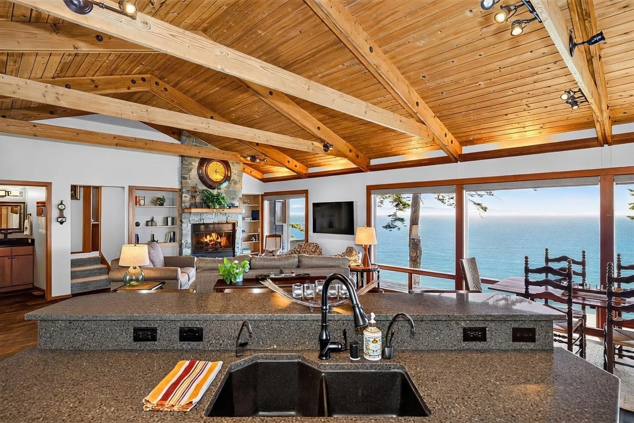 This perspective from the kitchen island looks out into an open-concept living area characterized by a vaulted wood-paneled ceiling with exposed beams. A stone-clad chimney breast serves as a focal point, while expansive windows provide a panoramic view of the ocean. The space features a warm, rustic aesthetic with comfortable seating and a seamless transition between the kitchen and the main living area.
