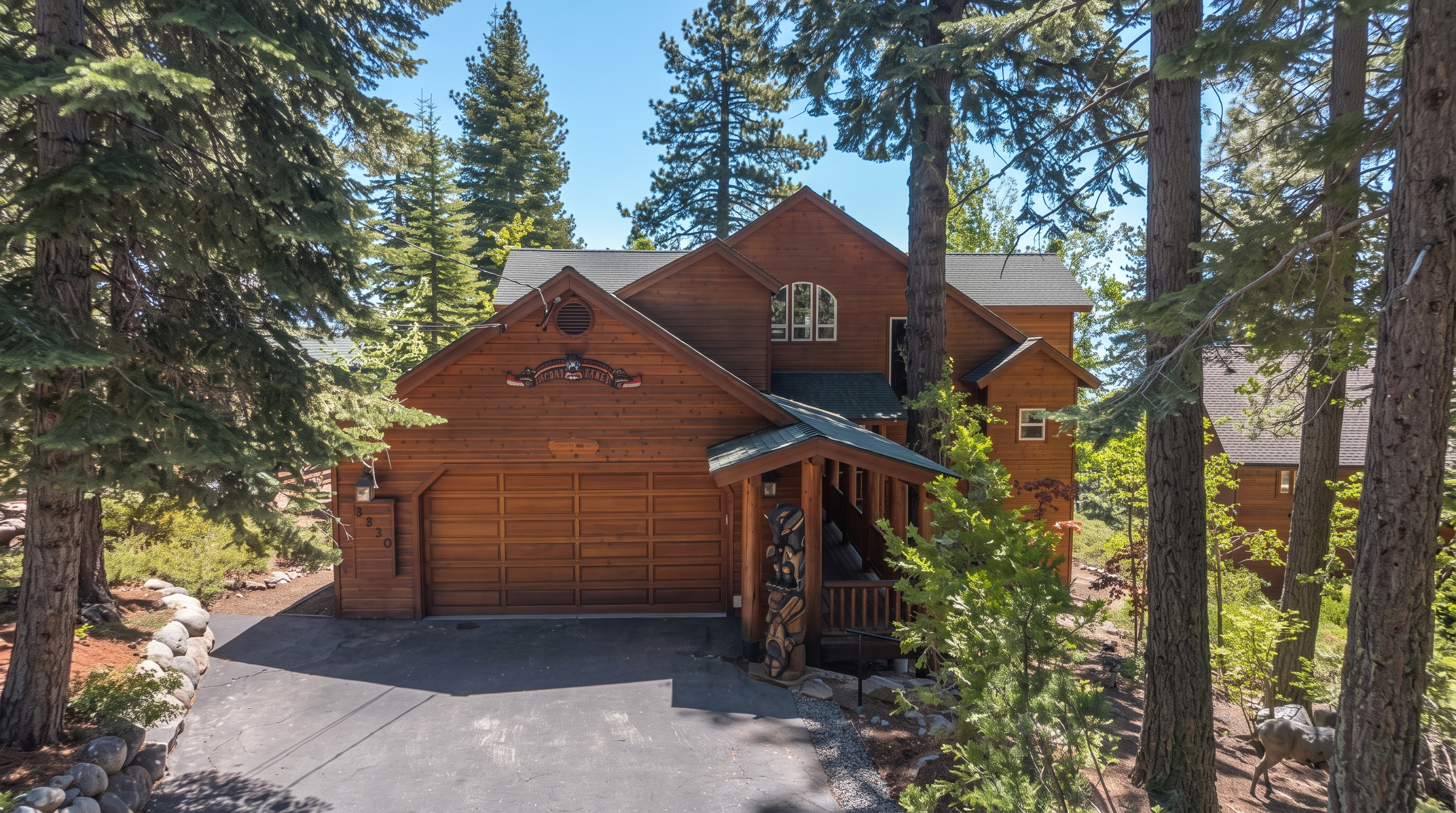 This charming, multi-level mountain cabin features warm-toned wooden siding, a large two-car garage, and an inviting covered porch adorned with a unique hand-carved wooden totem pole. The property is nestled in a lush, forested setting with mature pine trees framing the residence, creating a secluded and peaceful atmosphere. The architectural design includes gabled rooflines and classic shingle detailing, characteristic of a rustic woodland retreat.