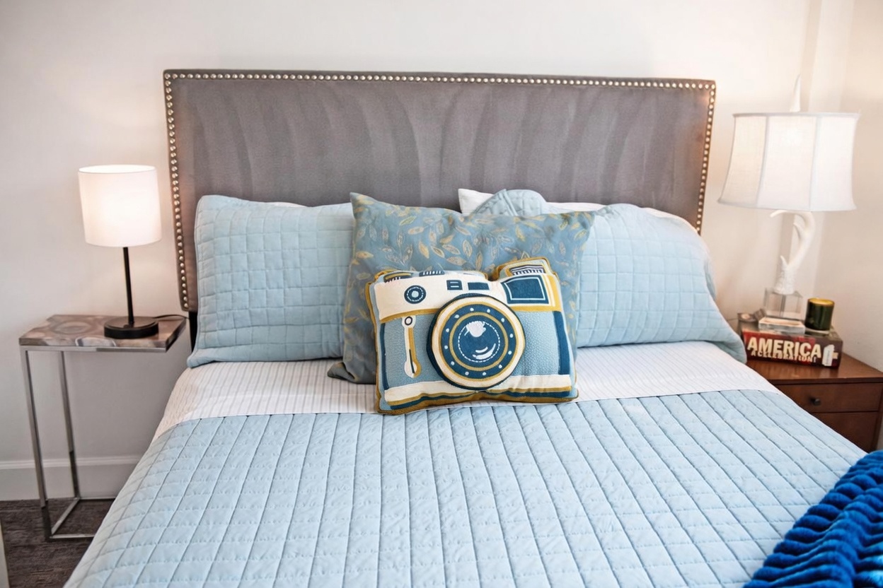 This cozy bedroom features a bed with a light blue quilted coverlet and a grey upholstered headboard accented with nailhead trim. The bed is styled with a decorative camera-shaped throw pillow and a patterned accent pillow, flanked by two bedside tables with matching lamps. The overall aesthetic is inviting and playful, creating a comfortable atmosphere for guests.
