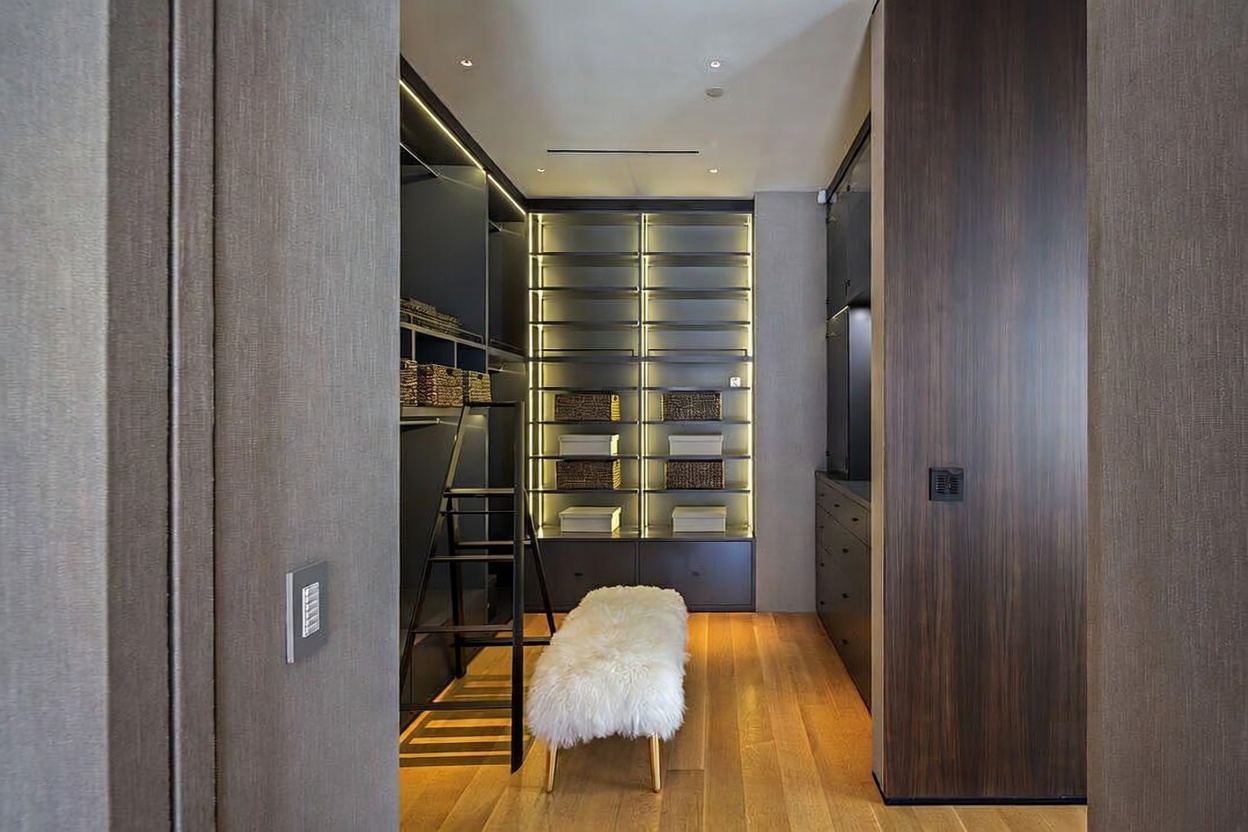 This sophisticated walk-in closet features dark wood cabinetry with integrated LED lighting that illuminates the shelving and storage sections. A plush, white faux-fur bench sits in the center of the room on light wood flooring, while a black rolling ladder provides access to higher storage areas. The perspective is framed through a doorway, highlighting the clean, modern aesthetic and organized layout of the space.