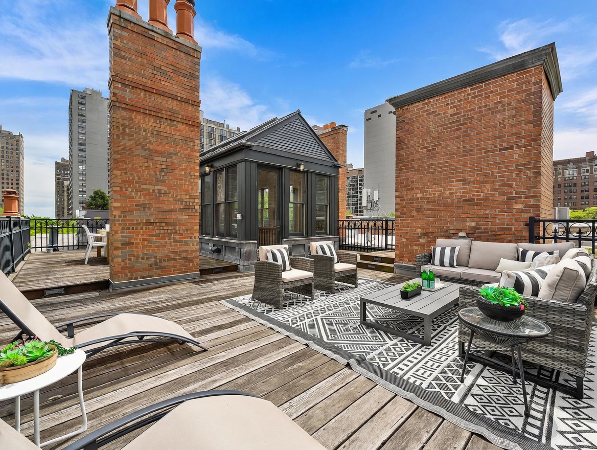 This expansive rooftop deck offers a sophisticated outdoor living space with a wooden plank floor and a central sunroom structure. The area is furnished with a comfortable sectional sofa, a coffee table, and lounge chairs, all arranged on a patterned area rug. Large brick chimneys frame the scene, providing a classic urban aesthetic against a clear blue sky.
