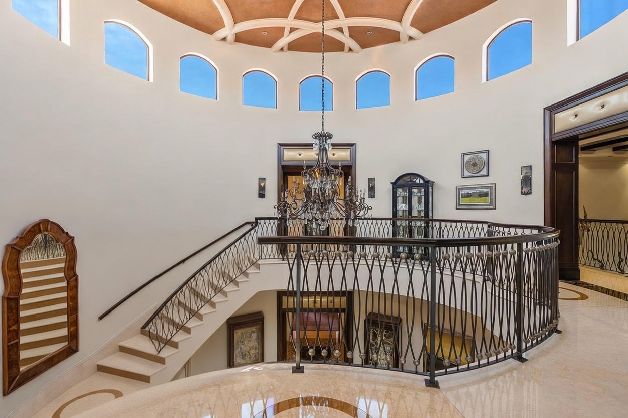 This grand foyer features a sweeping curved staircase with an ornate metal railing, overlooking a lower level. The space is defined by a dramatic rotunda ceiling with recessed lighting and a series of arched windows that flood the area with natural light. A large, intricate chandelier serves as the focal point, emphasizing the luxurious and architectural elegance of the home's entryway.