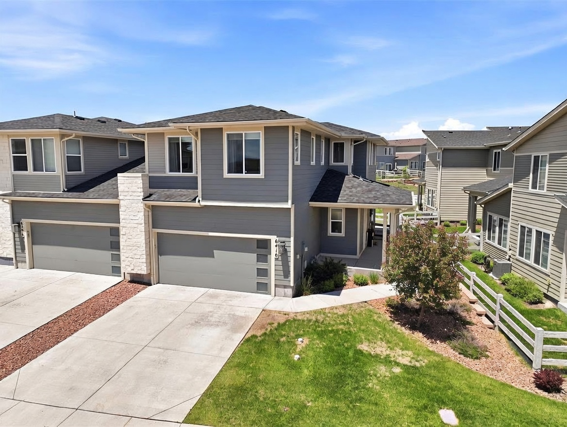This image presents an elevated, angled view of a two-story suburban residence featuring gray horizontal siding and a stone-clad chimney breast accenting the garage area. The home includes a multi-car garage, a covered front porch, and a well-maintained lawn with a white picket fence bordering the side yard. The perspective captures the architectural layout of the home in relation to the neighboring properties under a clear blue sky.
