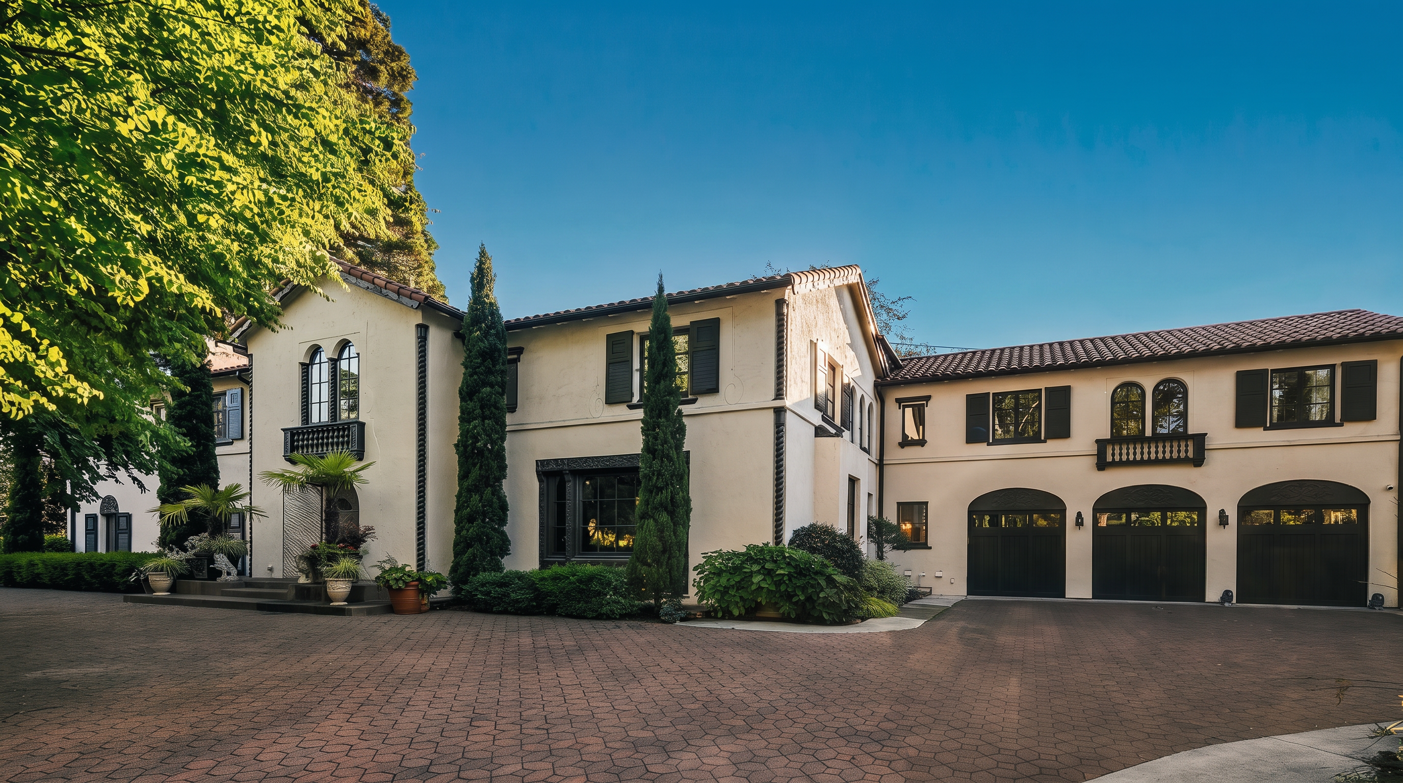 This elegant Mediterranean-style estate features a light-colored stucco facade with dark shutters and a multi-car garage. The property is framed by mature trees and manicured landscaping, with a spacious, patterned brick driveway leading to the main entrance. The architectural design includes arched garage doors and a decorative balcony, creating a sophisticated and welcoming curb appeal.
