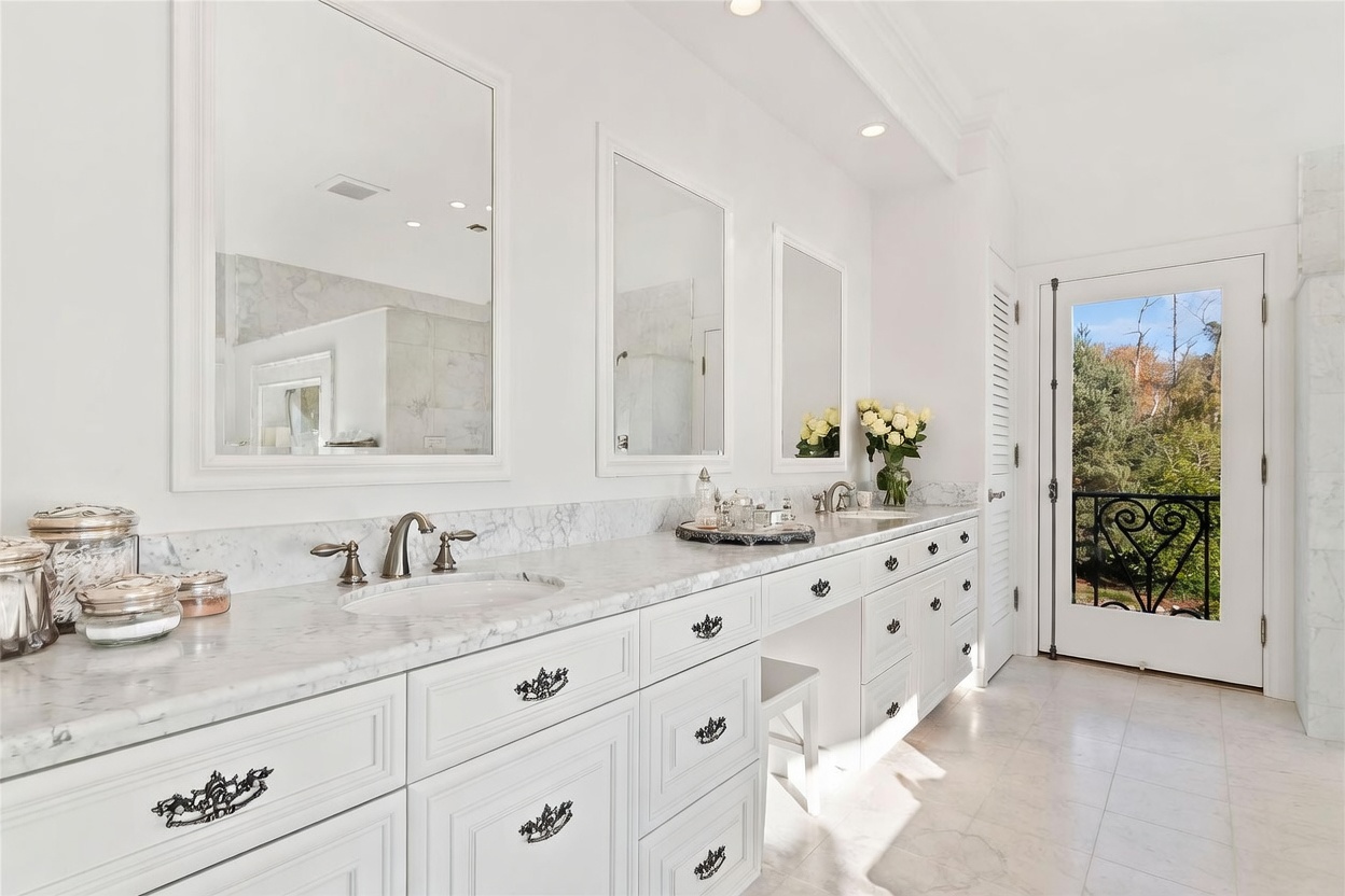 This bright and elegant primary bathroom features a long vanity with white cabinetry, dark hardware, and a light-colored stone countertop. Three separate mirrors are mounted above the vanity, which includes a dedicated makeup seating area, while a glass door leads to an outdoor balcony. The space is finished with light-toned tile flooring and recessed lighting, creating a clean and sophisticated atmosphere.