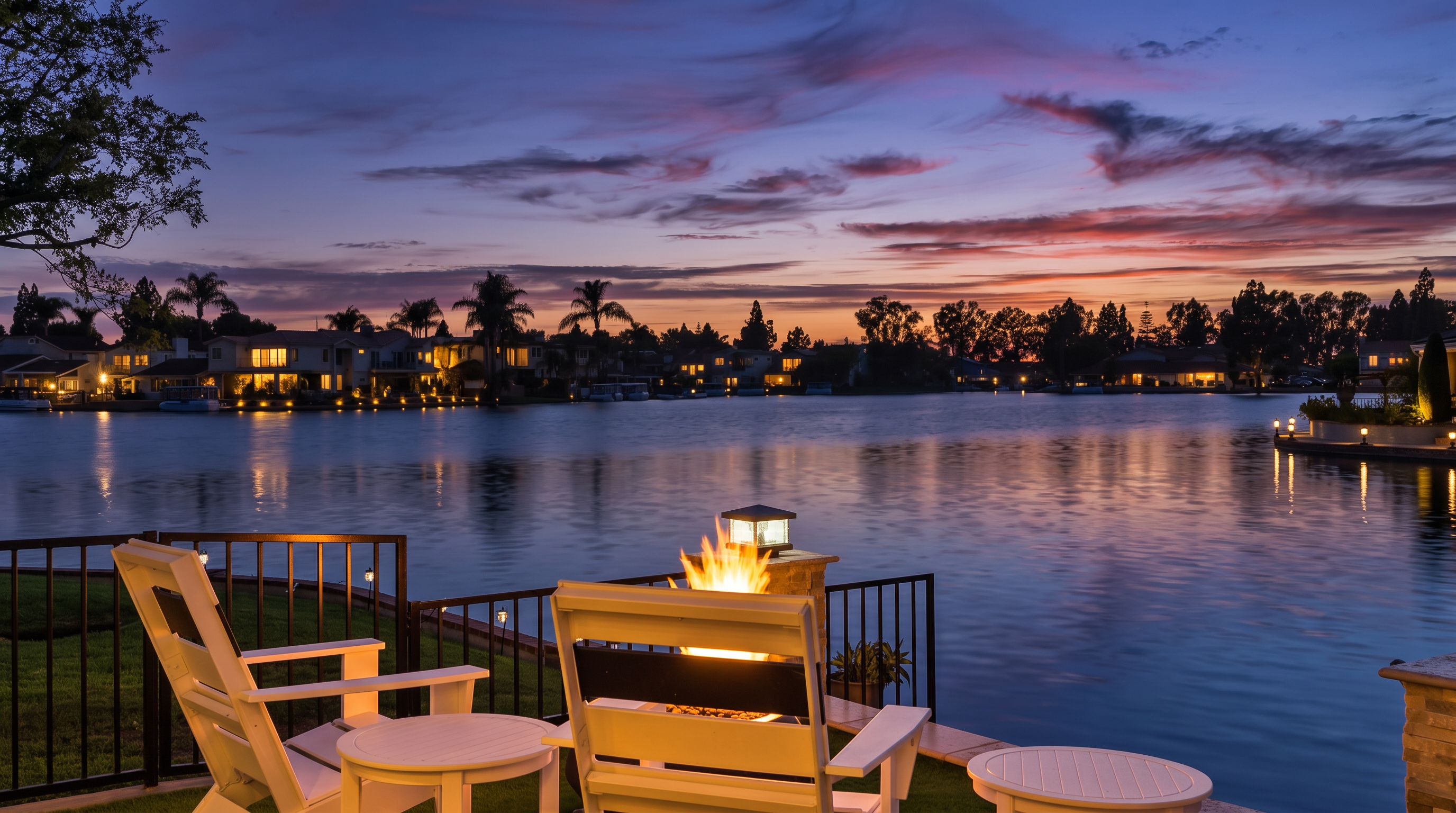 This serene waterfront patio features white Adirondack-style chairs and side tables arranged around a glowing fire pit. The space offers a direct, picturesque view of a peaceful lake at sunset, framed by lush landscaping and residential homes across the water. It creates an inviting and tranquil atmosphere perfect for evening relaxation or outdoor entertaining.